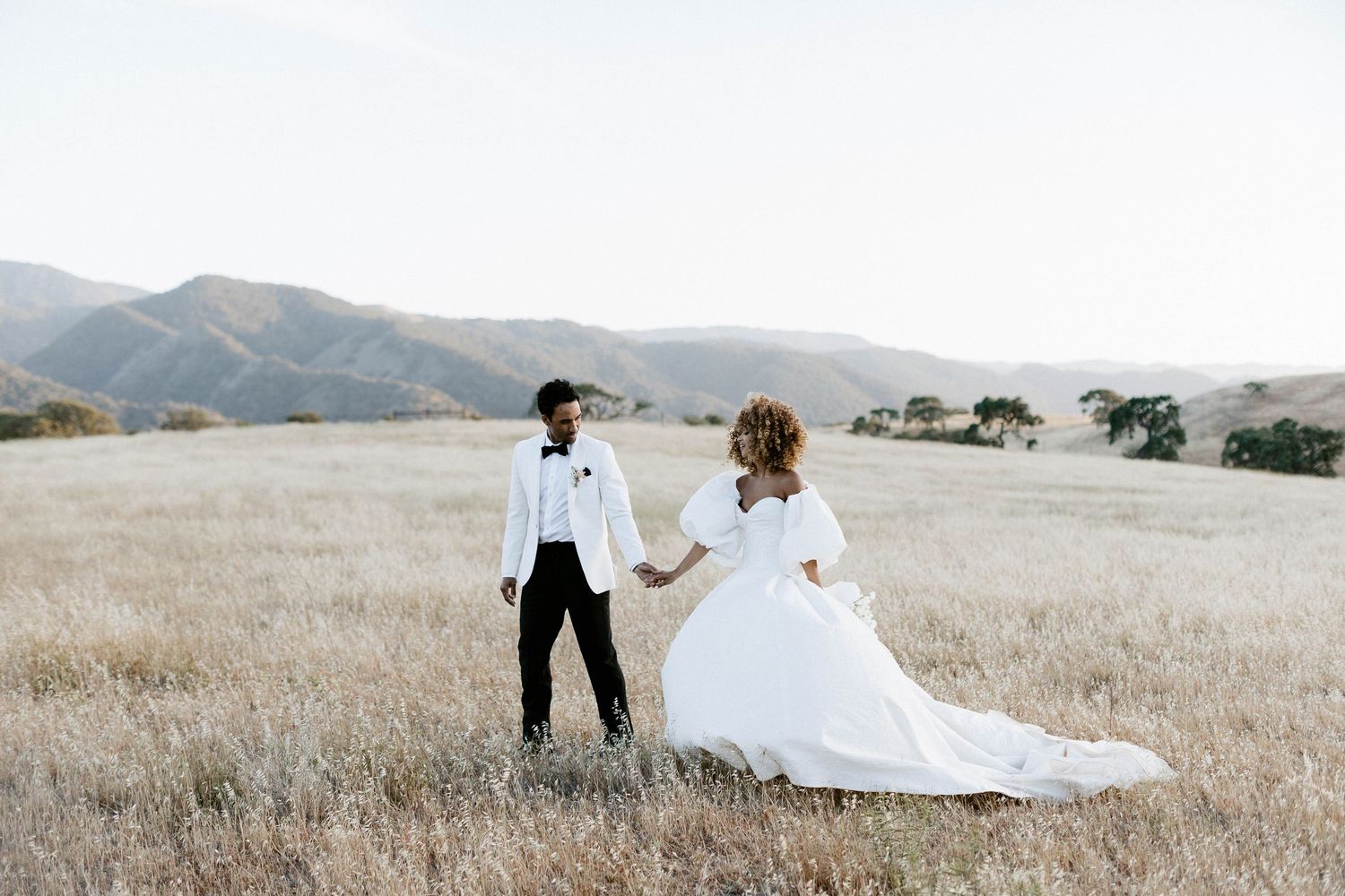A couple in wedding attire holds hands while standing in a golden field with Santa Barbara mountains in the background in Kestrel Park Cali.