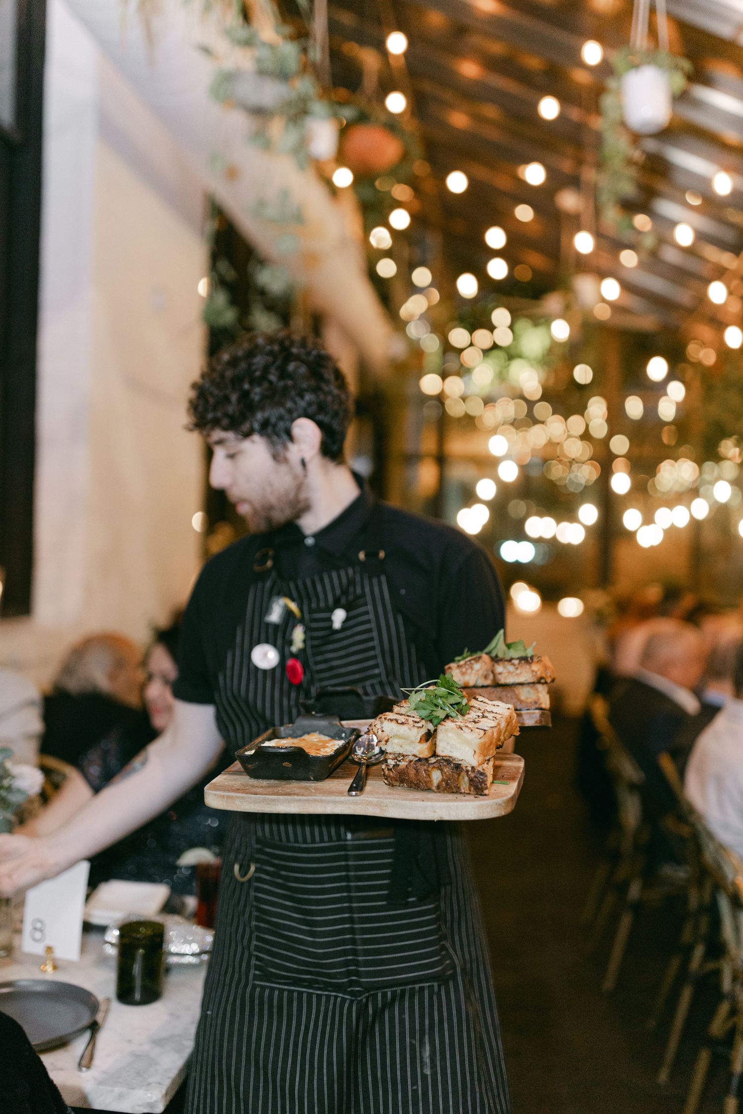 A server in striped apron carries plates of food through a dimly lit restaurant with string lights.