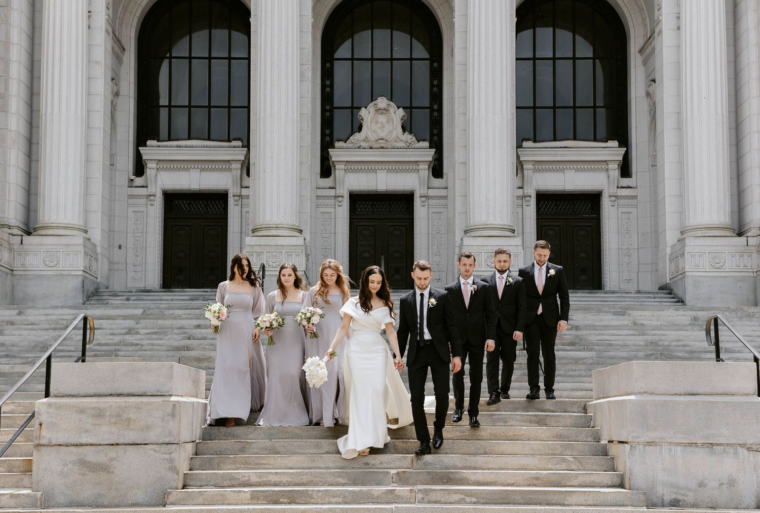 Wedding party members in black and white attire descending grand stone steps of a classical building.