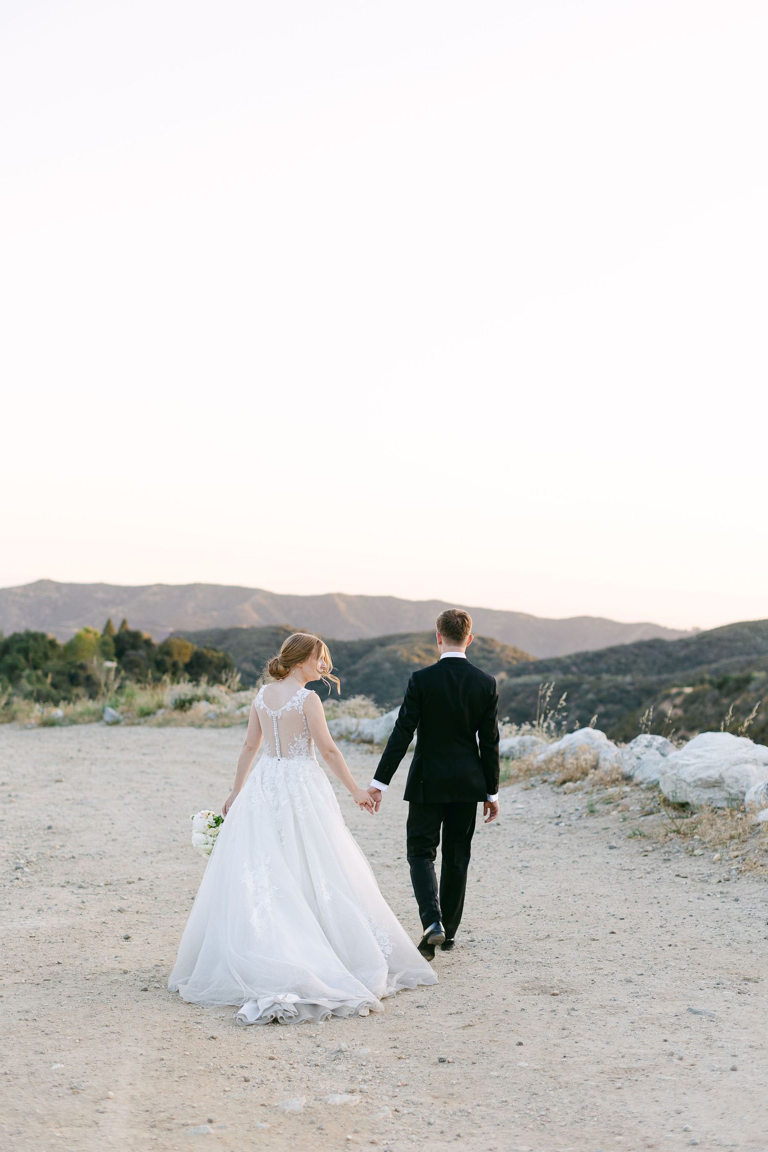 A couple in formal wedding attire walks along a desert path with mountains in the background.
