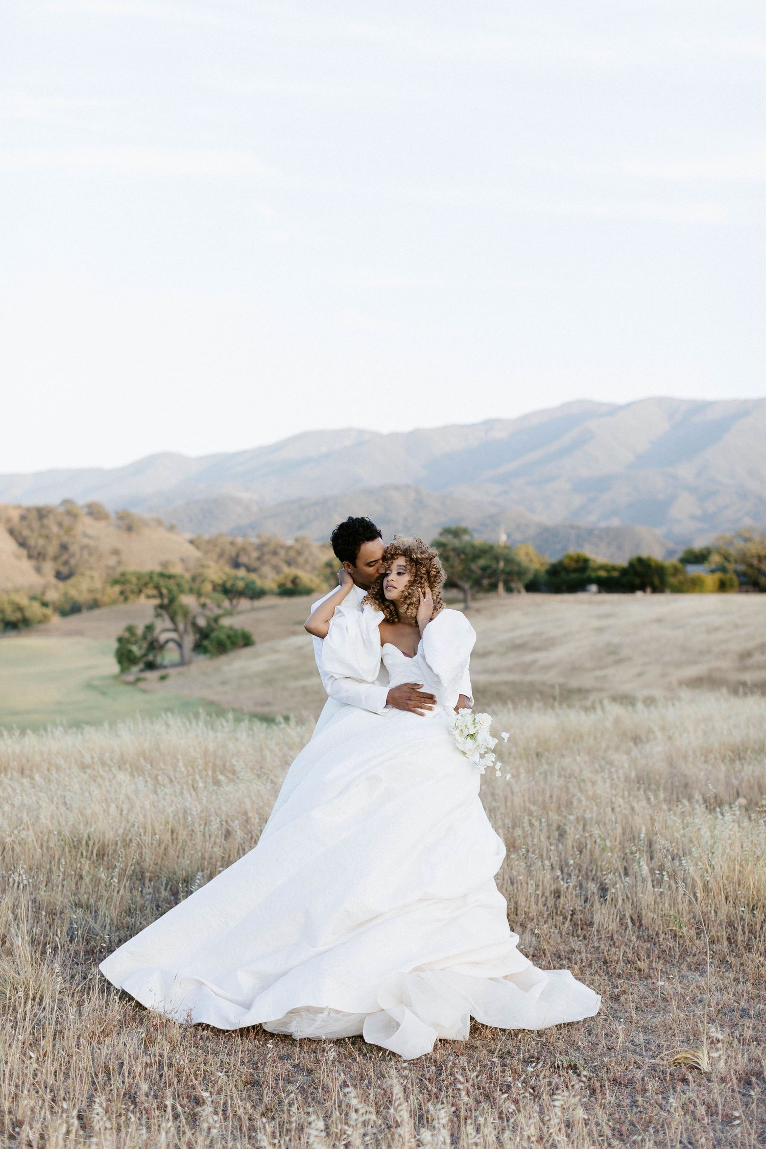 Newlyweds share an intimate moment in a rustic outdoor setting with rolling hills behind them.