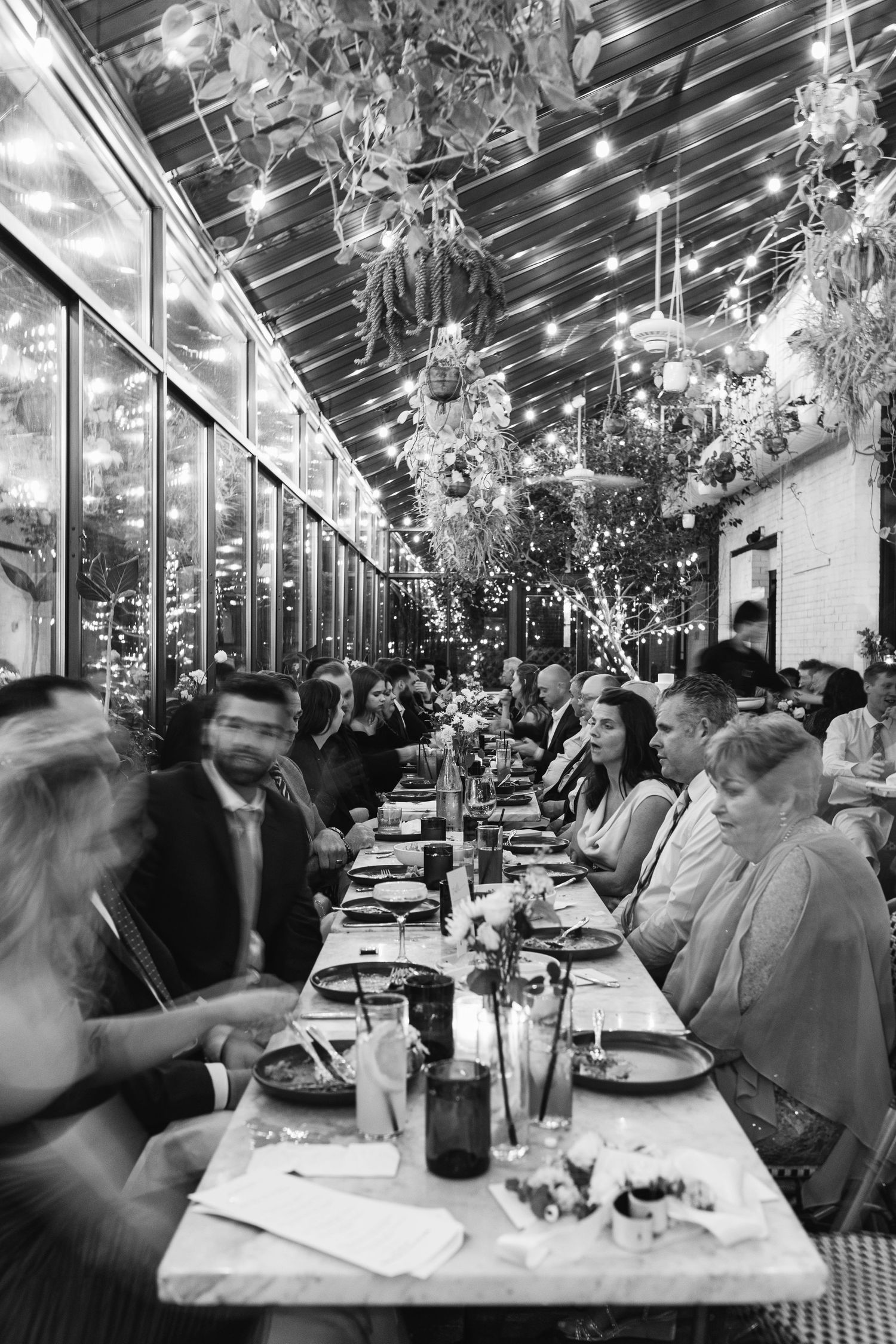 A black and white photo of wedding guests dining at a long table under crystal chandeliers.