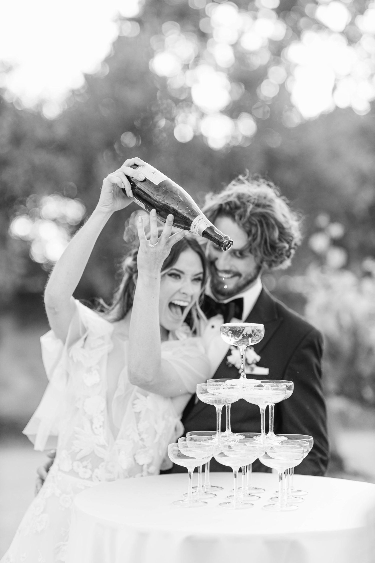 Wedding couple laughs joyfully while pouring champagne into a tower of glasses at an outdoor celebration.