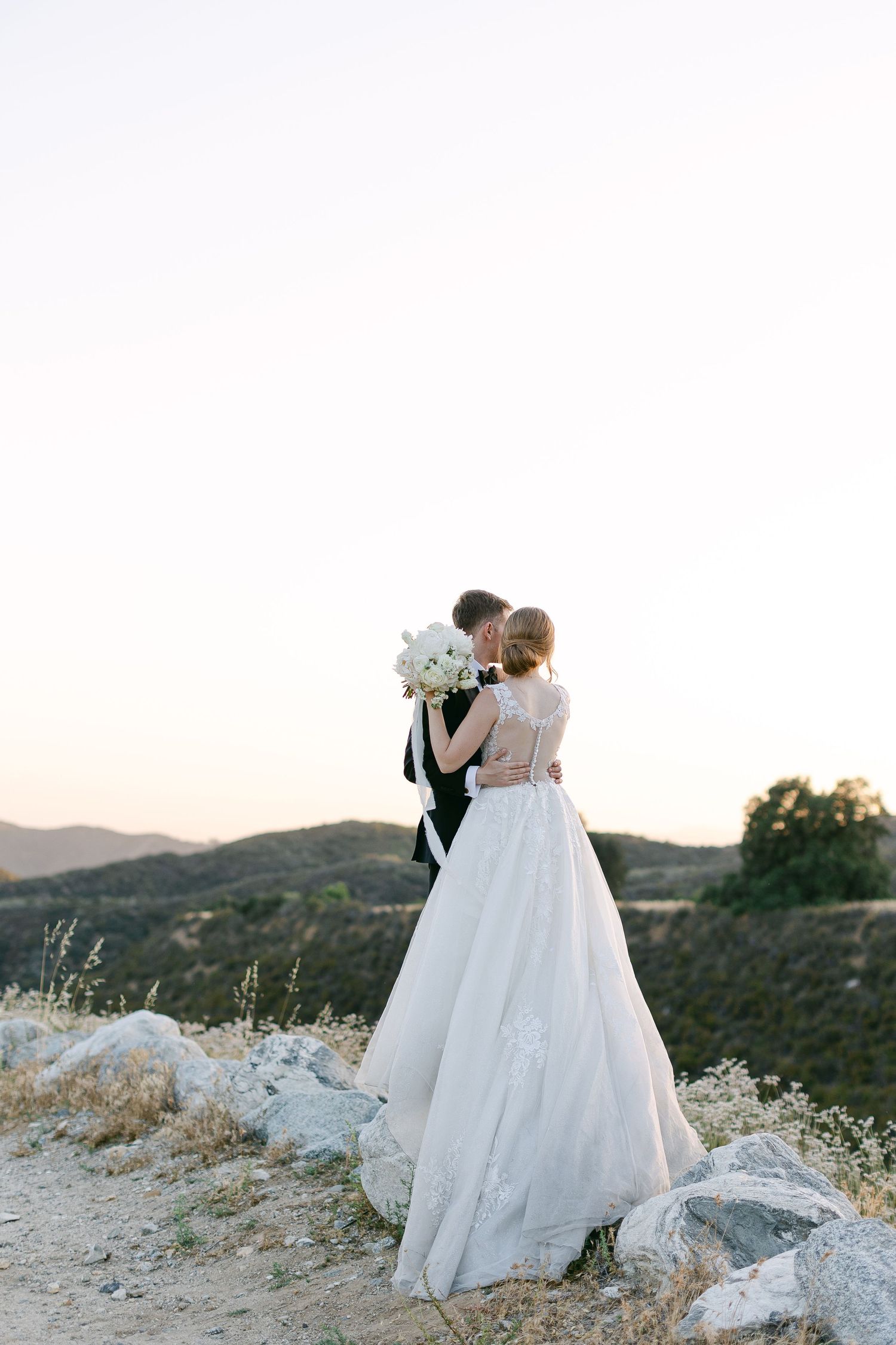 A couple in wedding attire stands together on a hilltop during golden hour.
