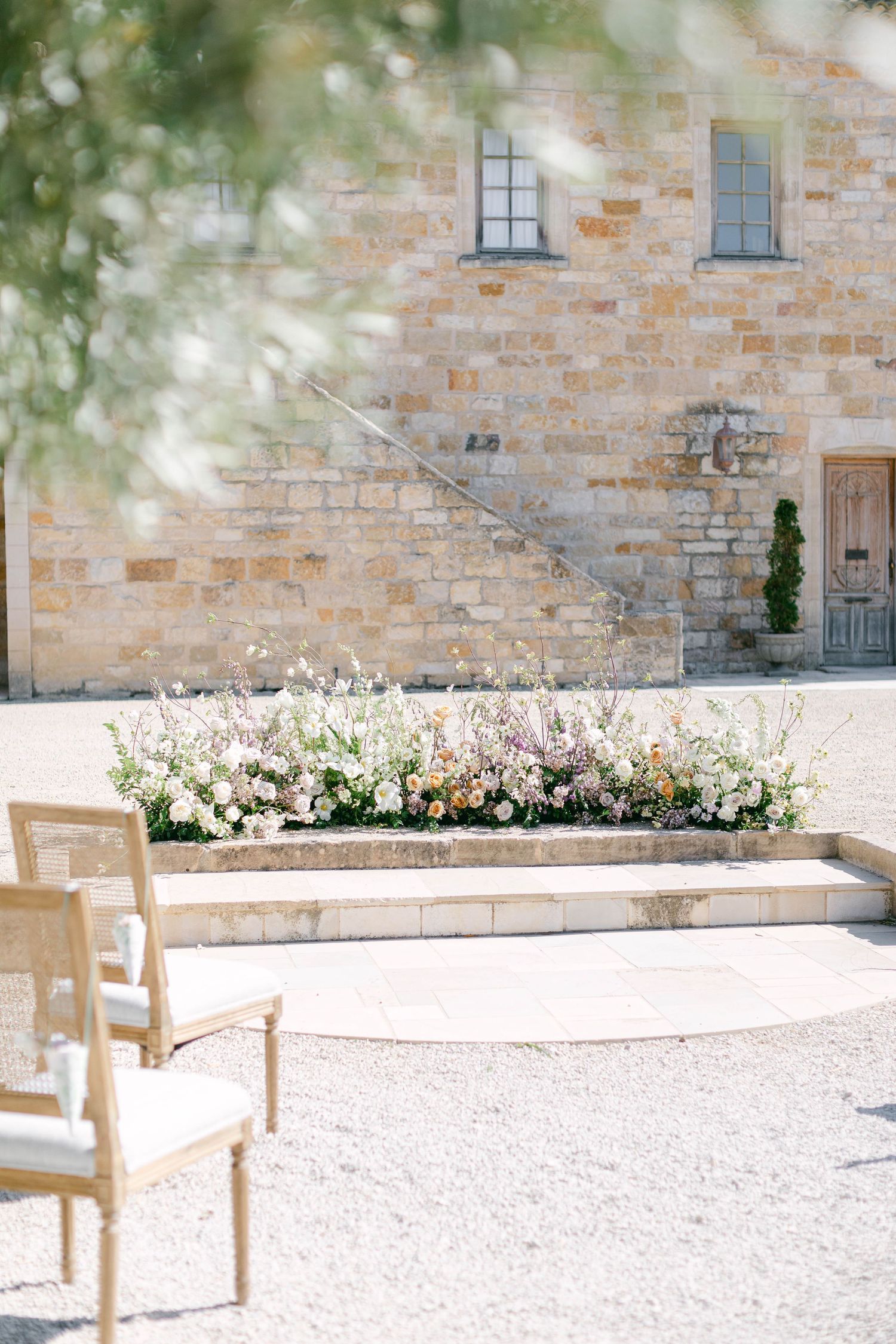 Outdoor wedding ceremony setup with wooden chairs and floral arrangements against stone building.