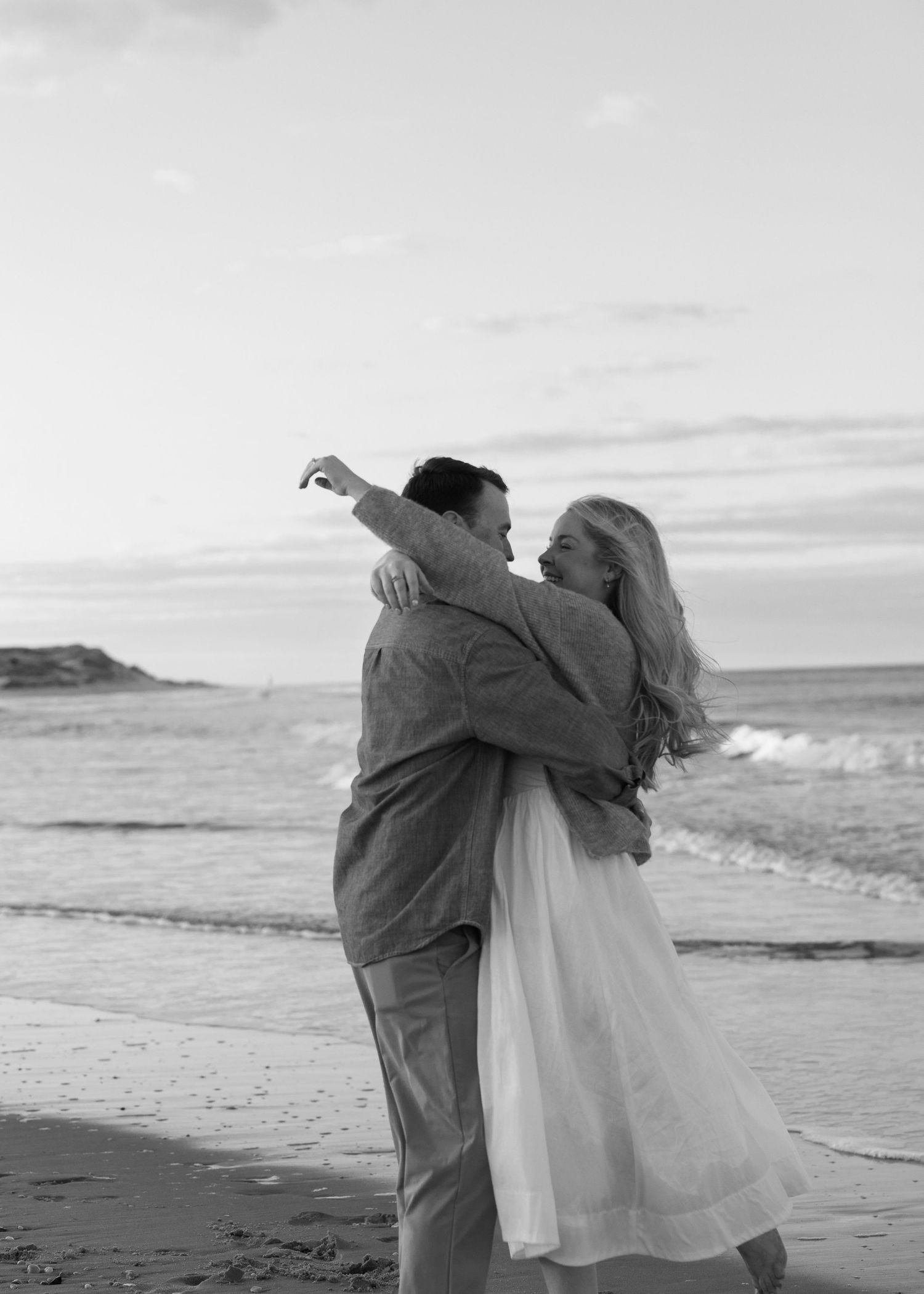 A couple dances together on a scenic Delaware beach for engagement photographs with waves crashing in the background in black and white.