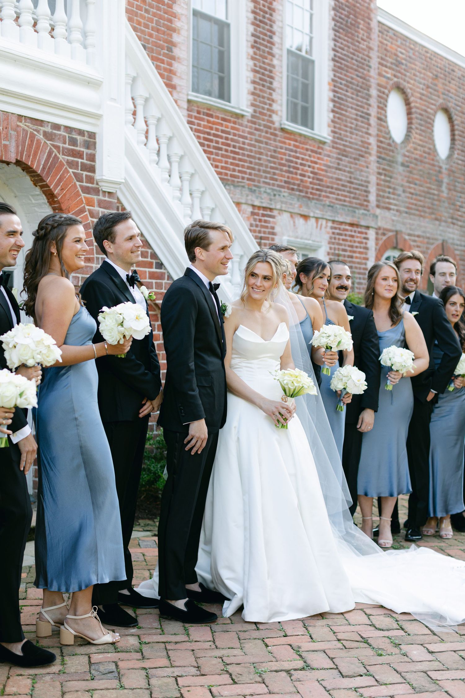 Wedding party in formal attire poses on brick pathway with bridesmaids in blue dresses and groomsmen in black suits.