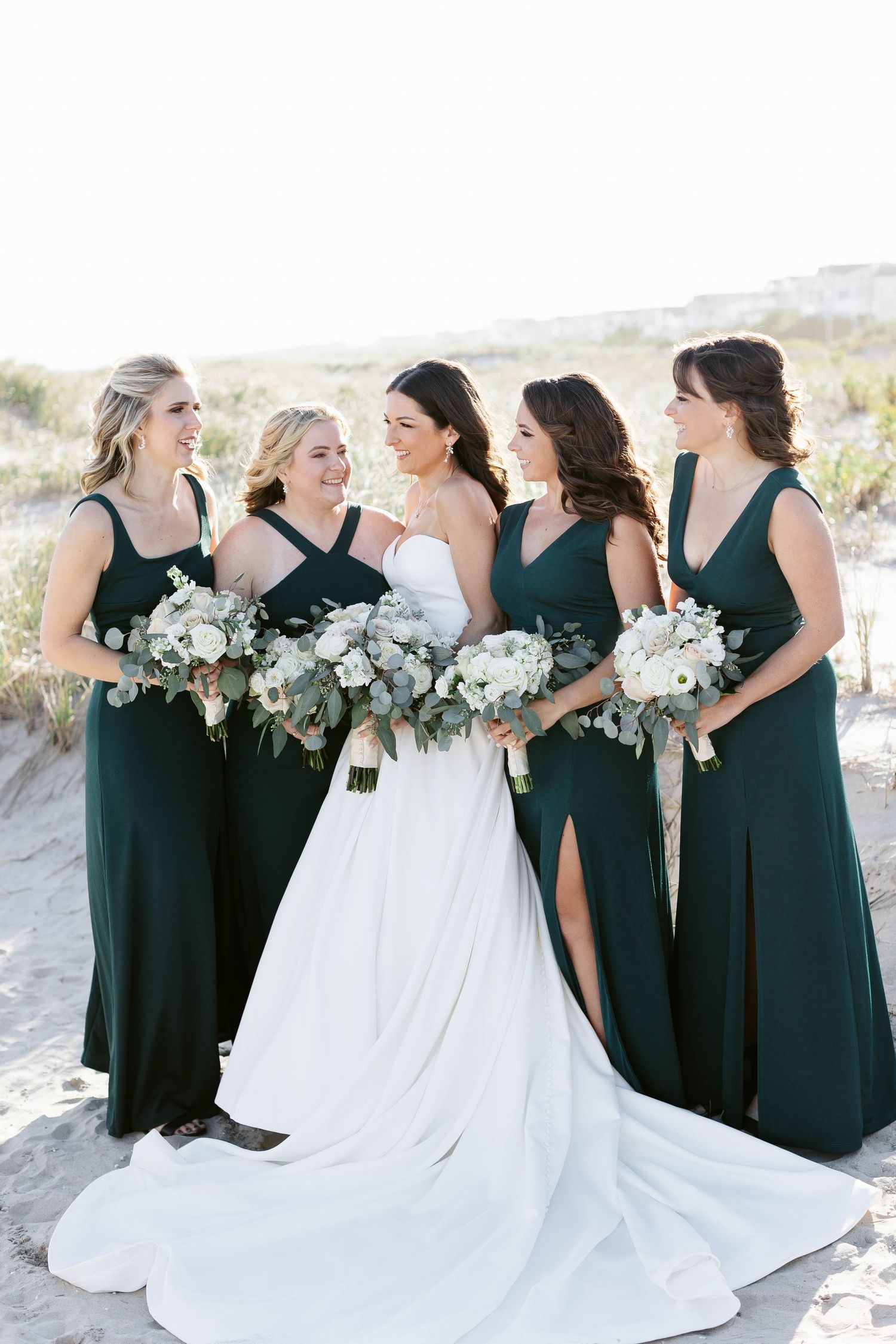 Bridesmaids in elegant dark green dresses pose with white bouquets on a beach alongside bride in flowing white gown.