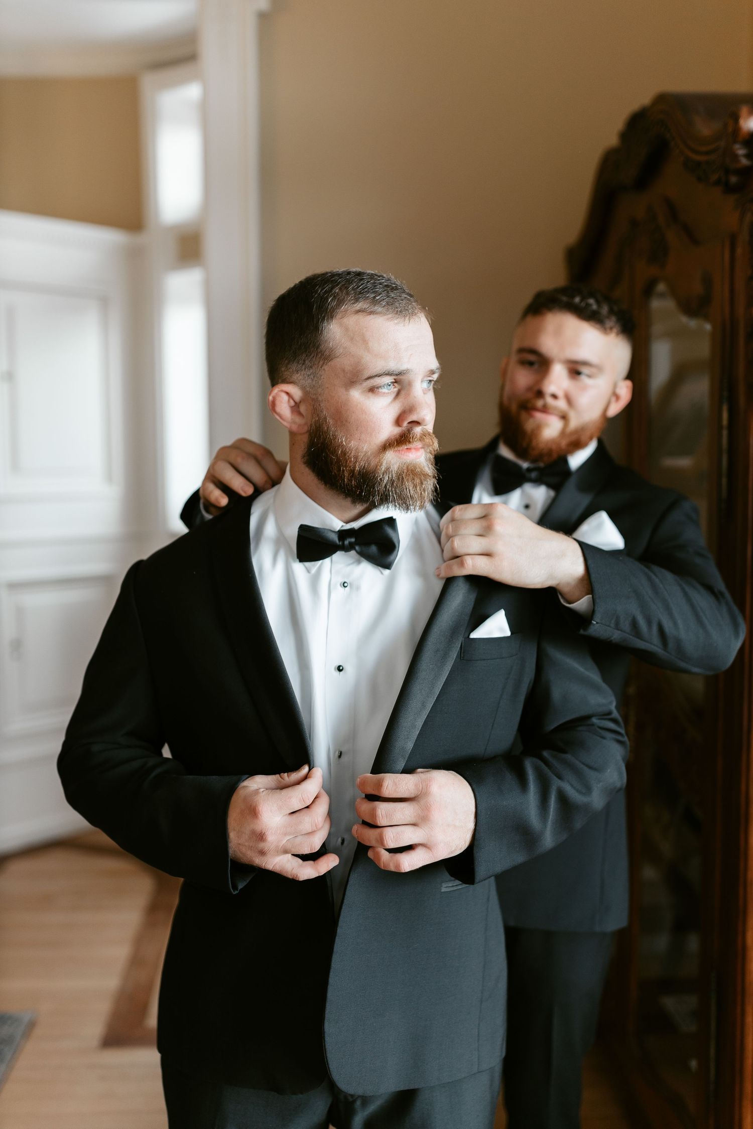 Two men in tuxedos adjust formal attire while getting ready.