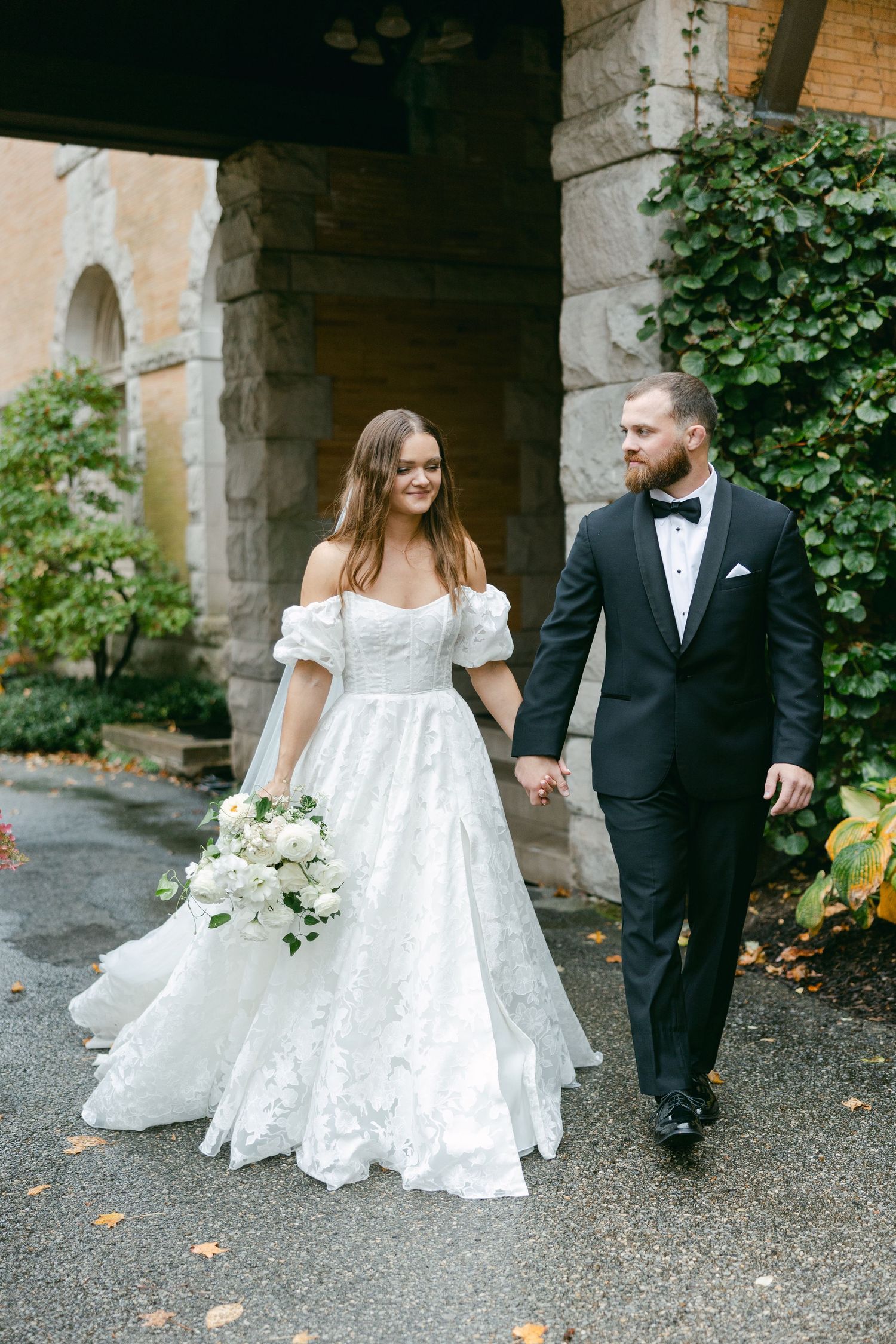 A wedding couple walks hand in hand under a stone archway, with the bride wearing an off-shoulder white gown.