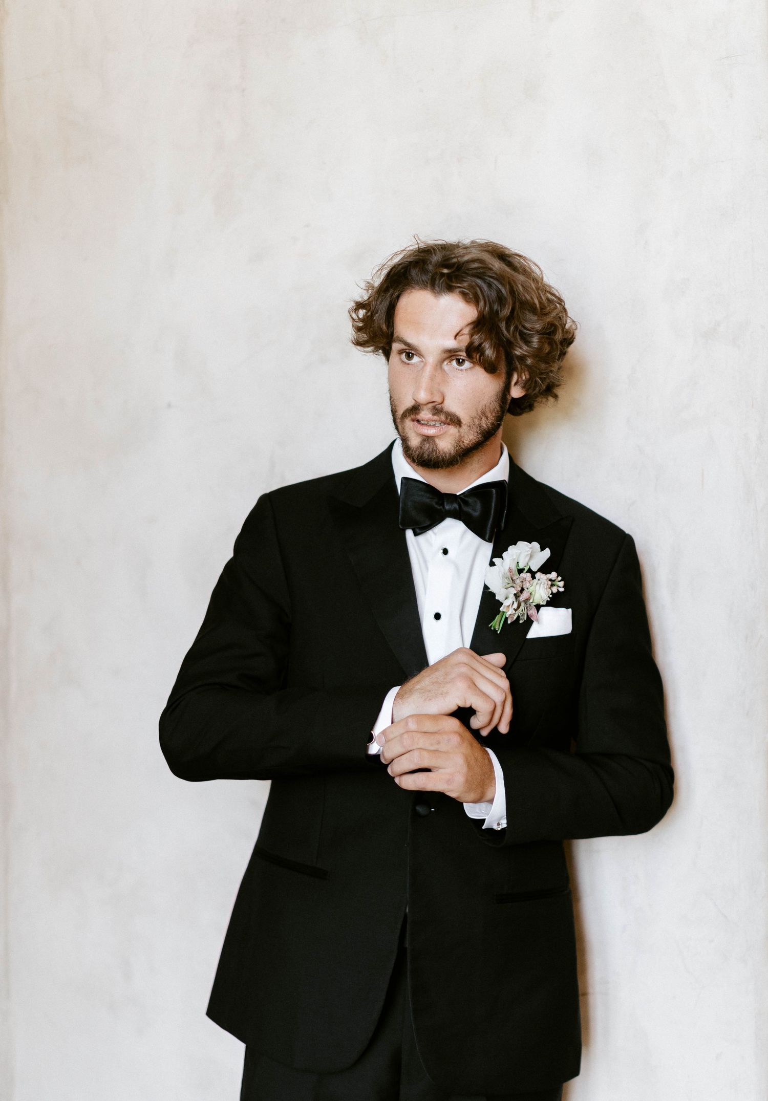 Groom in classic black tuxedo poses against white backdrop with boutonniere.