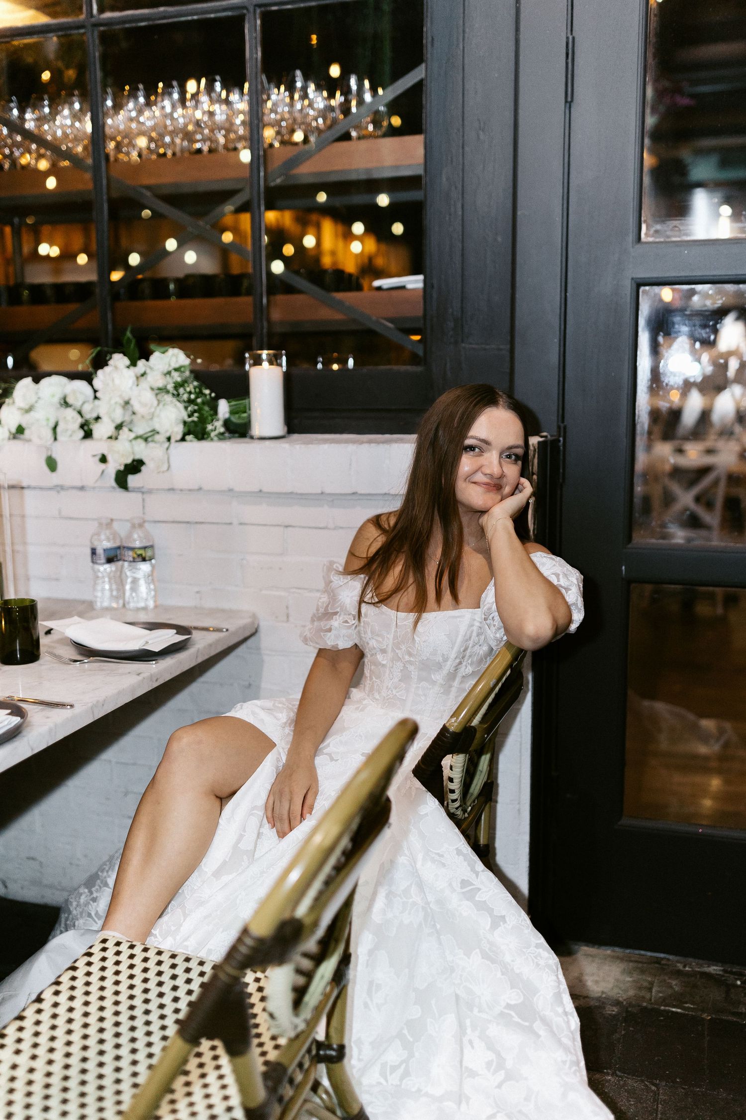 A bride in a lace wedding dress sits casually at a restaurant booth with candlelit ambiance.