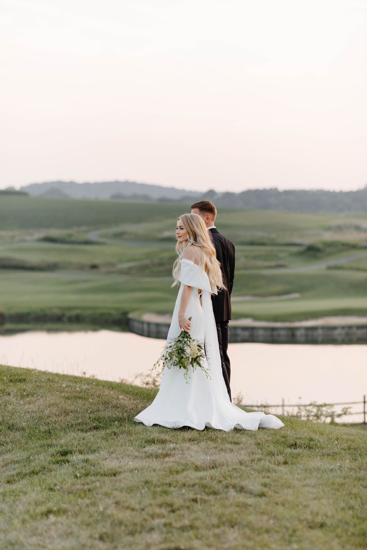 Wedding couple embraces on a hilltop overlooking a serene lake at sunset.