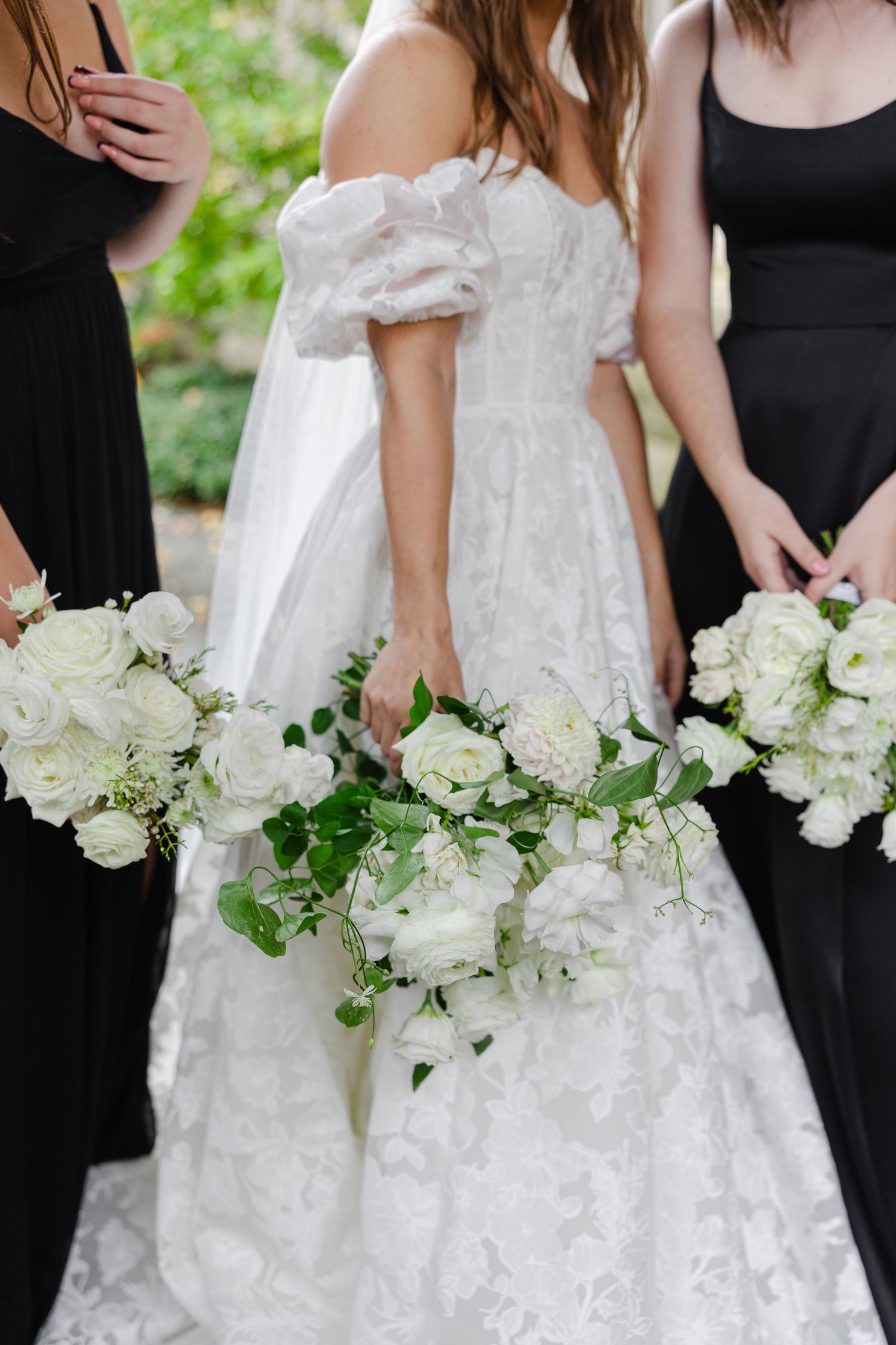 Bridesmaids in black dresses holding white rose bouquets next to bride in lace gown.