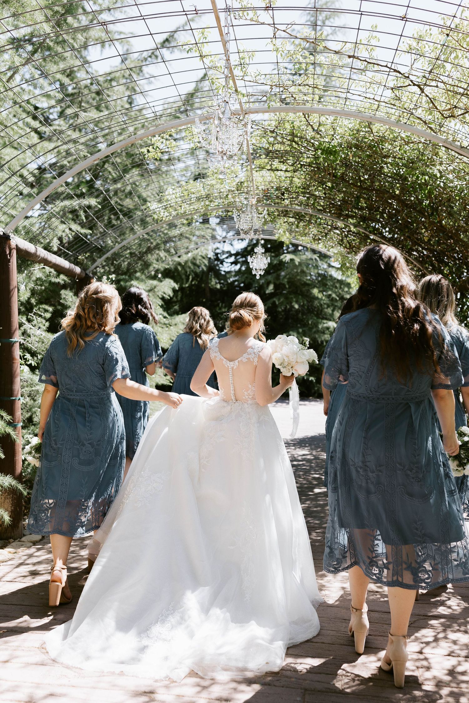 Bridesmaids in blue dresses walking with the bride under a greenhouse structure.
