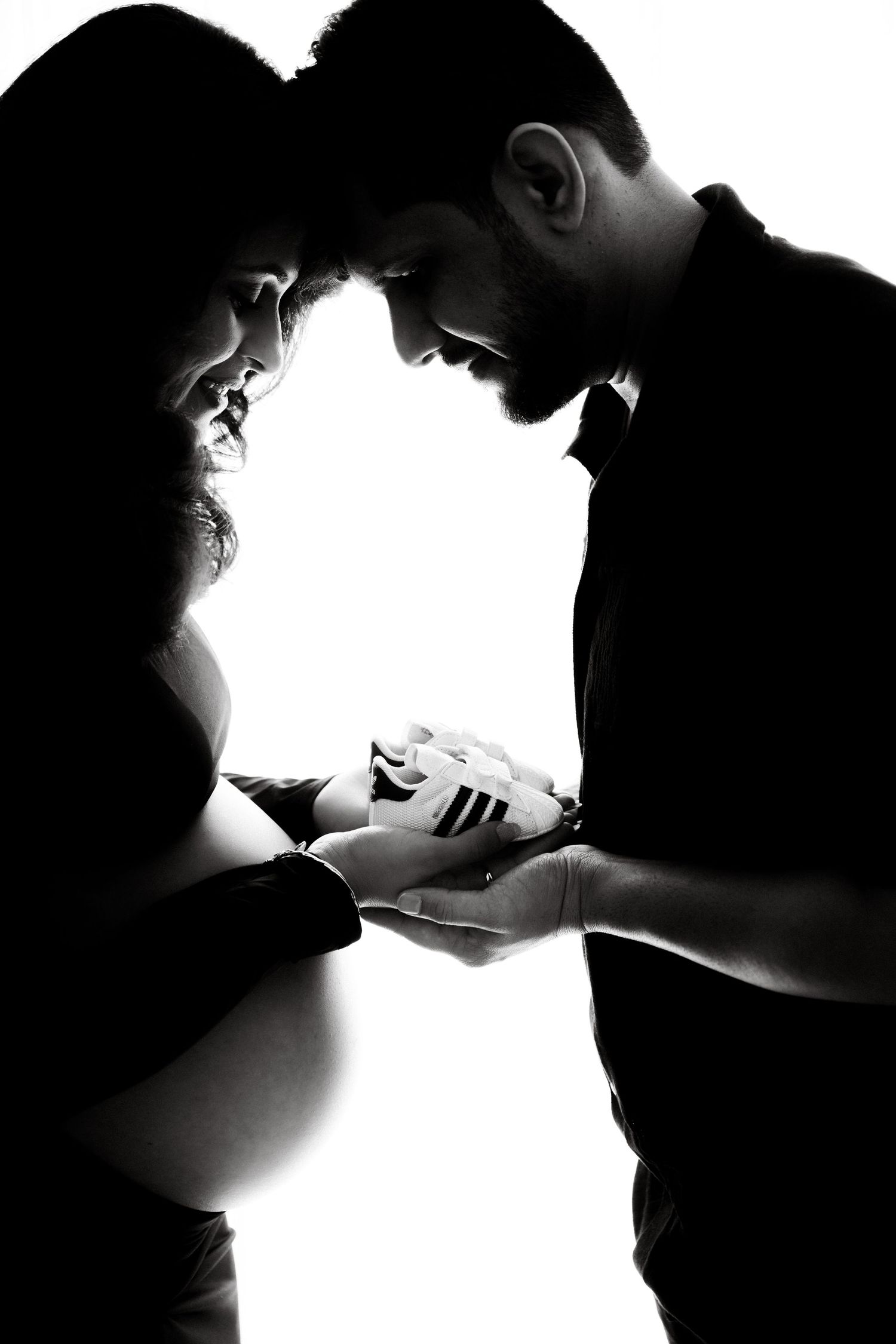 Black and white silhouette of pregnant couple holding tiny baby shoes, touching foreheads tenderly.