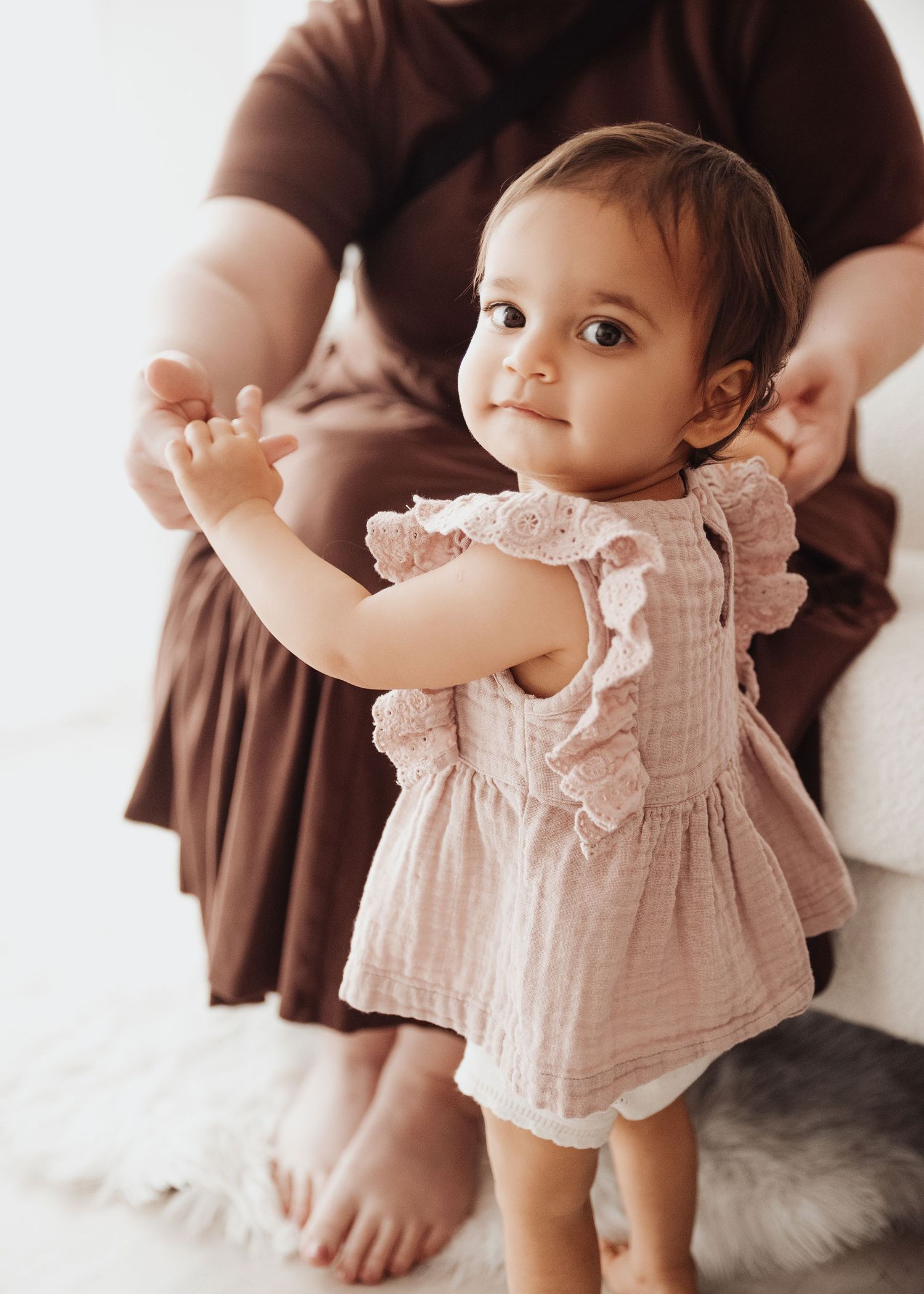 Toddler in a blush pink ruffled muslin dress stands holding an adult's hands on a soft white rug in a bright, airy room.