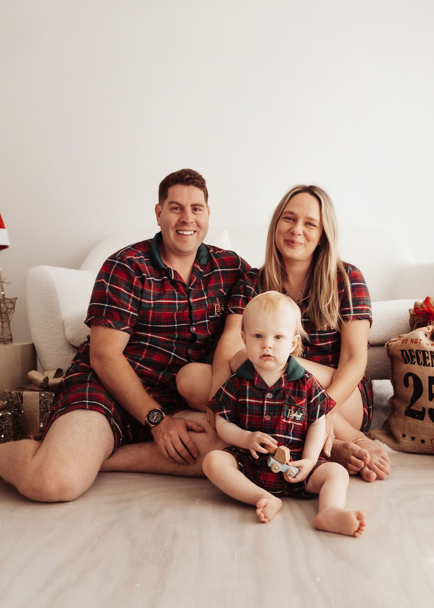 Smiling family of three in matching red plaid Christmas pajamas sitting together on a light floor beside holiday gifts and a burlap sack.