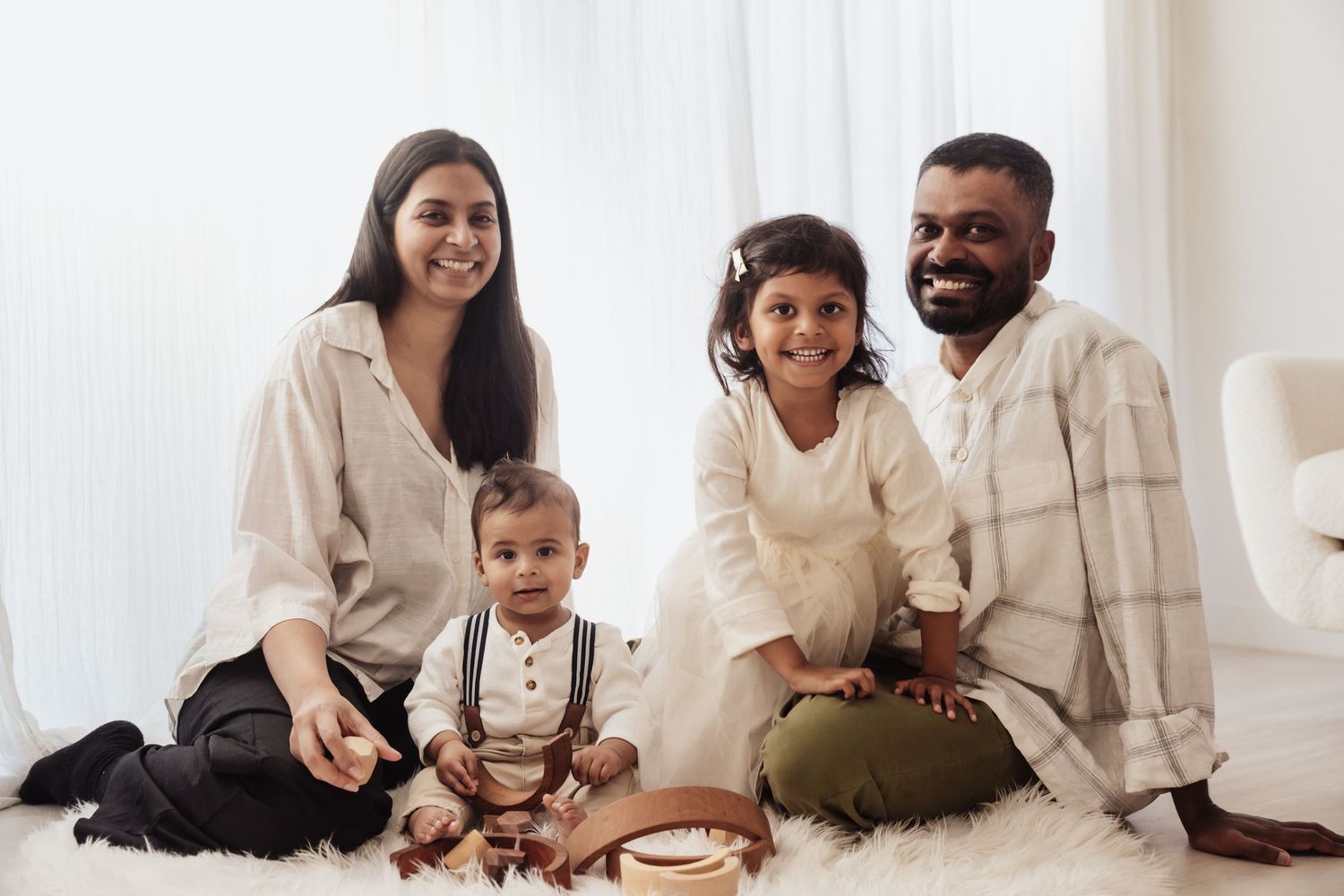 Smiling family of four in neutral linen outfits sitting together on a soft white rug in a bright, airy room with a toddler in suspenders.