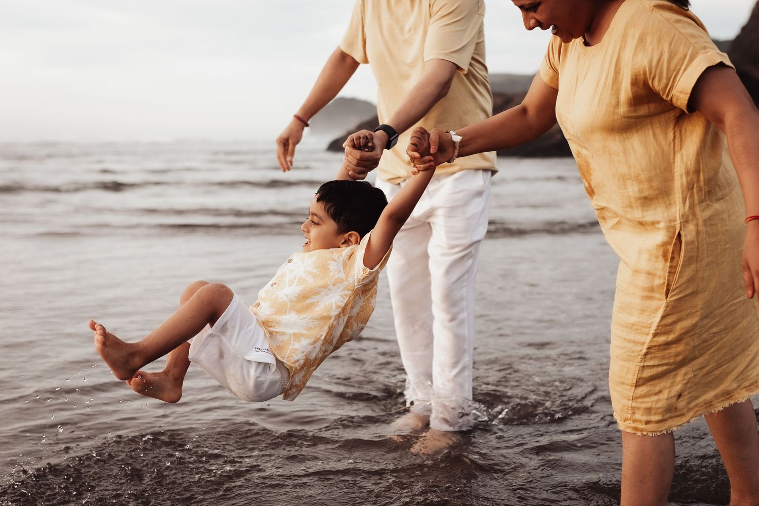 Family swinging child at beach, parents holding hands of happy kid over ocean waves at sunset.