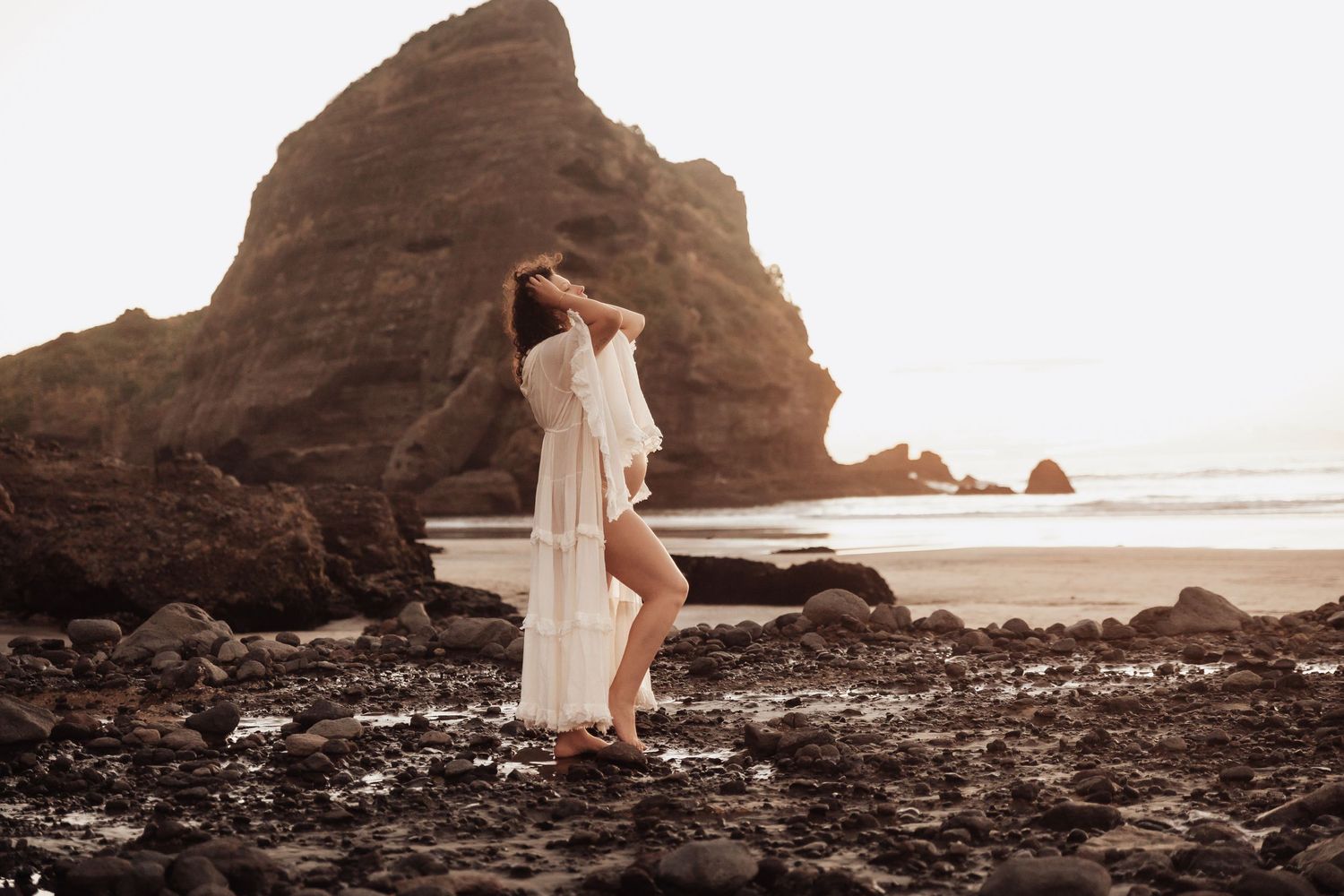 Woman in white sheer dress poses on rocky beach with dramatic sea stacks at sunset.