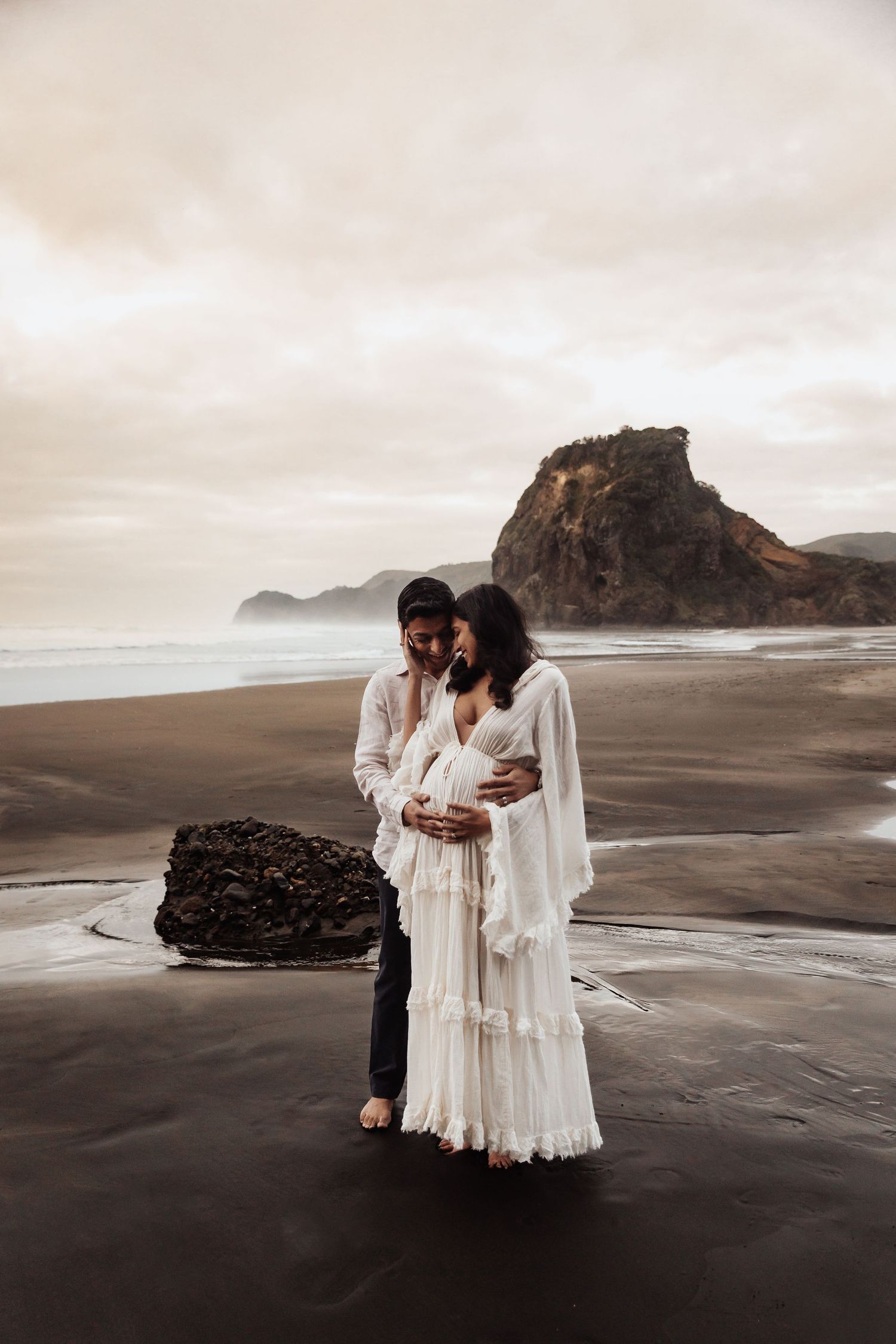 Couple in white embracing on black sand beach with dramatic rock formation and moody sky backdrop.
