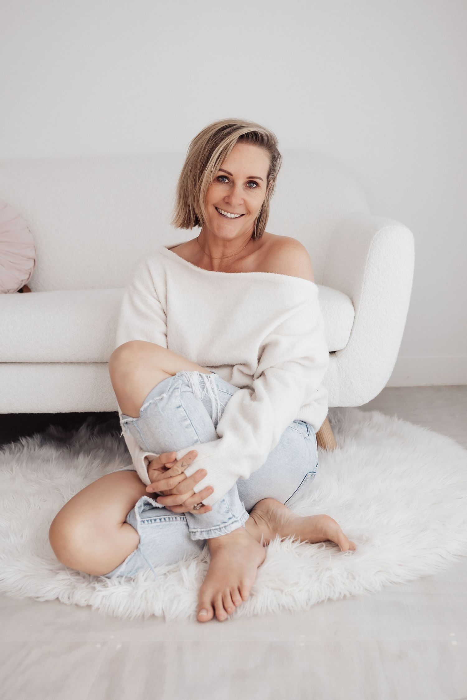 Smiling blonde woman sitting on a fluffy white rug in a cozy living room, wearing an off-shoulder sweater and ripped jeans.