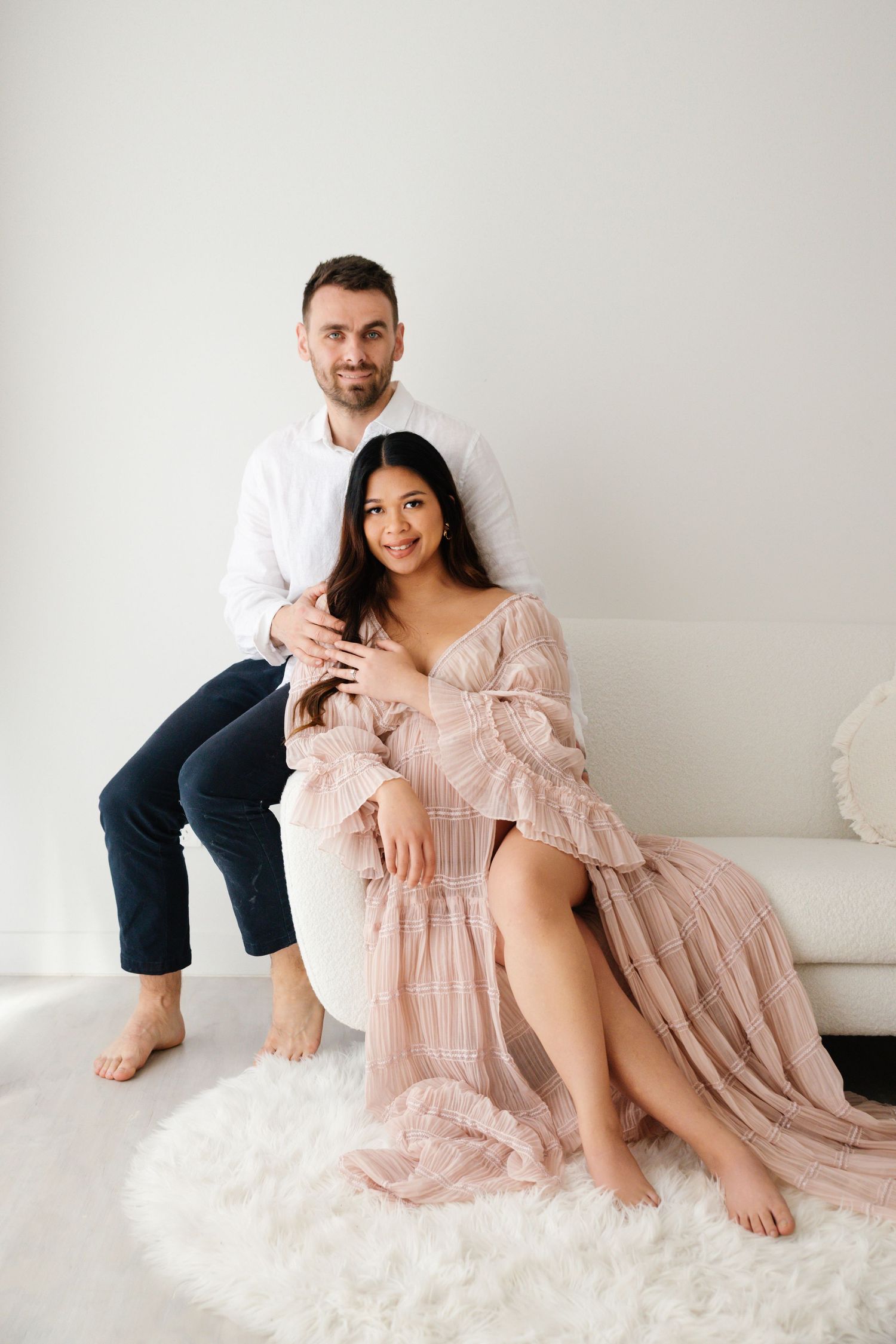 Couple posing for maternity photo shoot, woman in pink flowy dress seated on white fur rug, man in white shirt behind her.