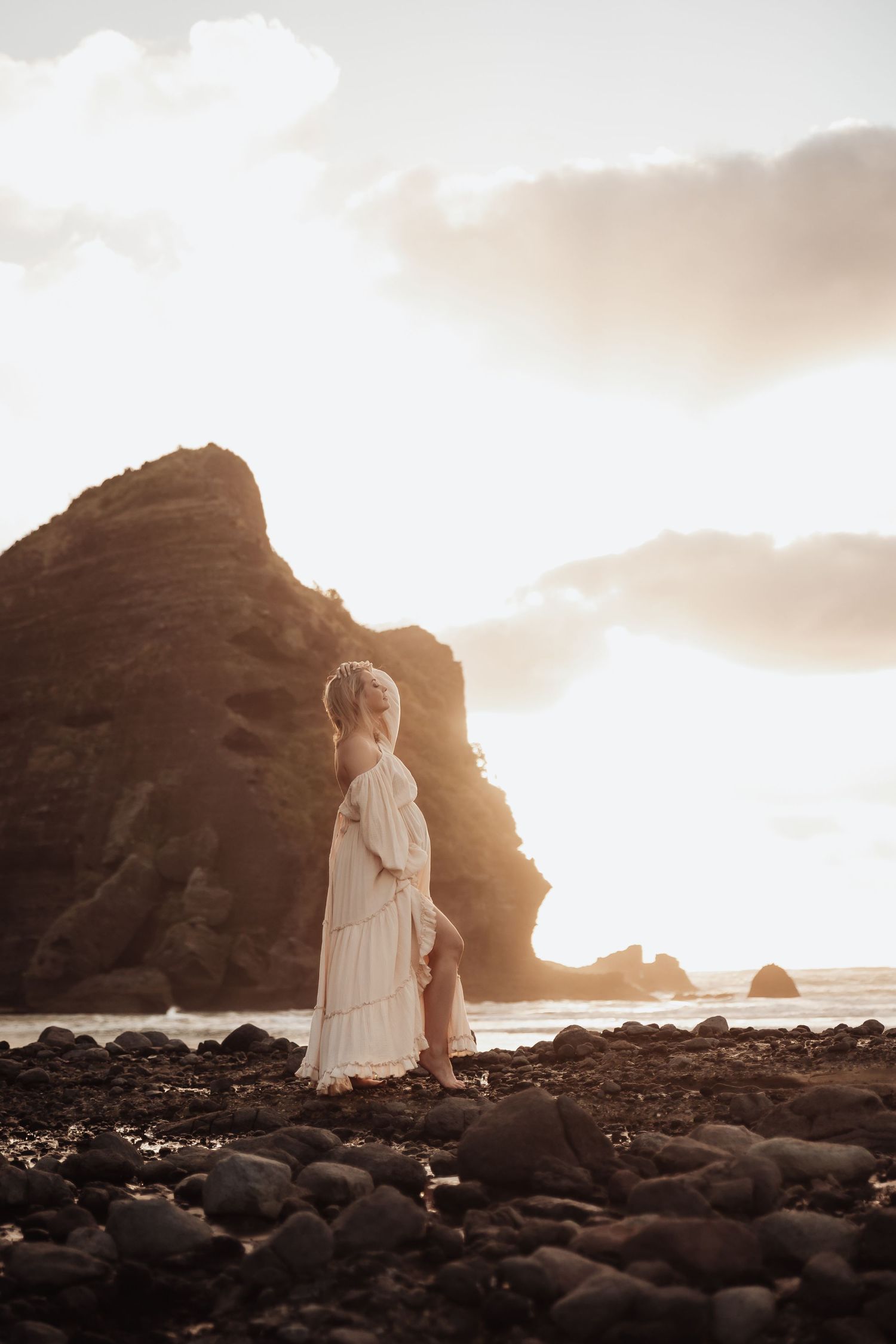 Woman in white dress standing on rocky beach at golden sunset with dramatic ocean cliffs in background.