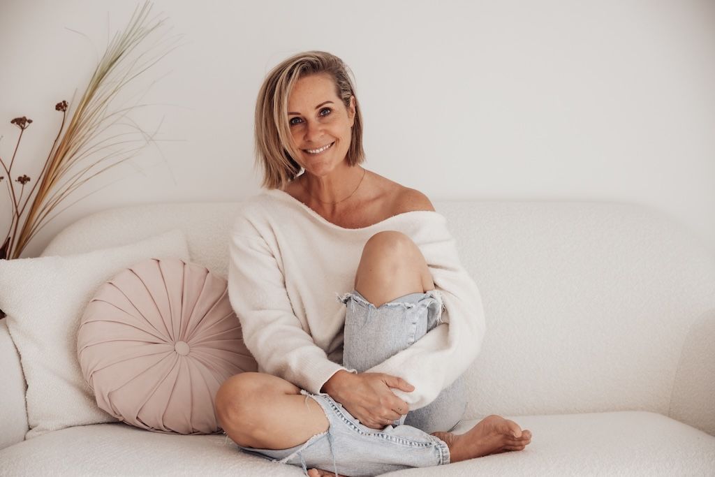 Smiling blonde woman sitting cross-legged on a white sofa wearing an off-shoulder sweater and ripped jeans.
