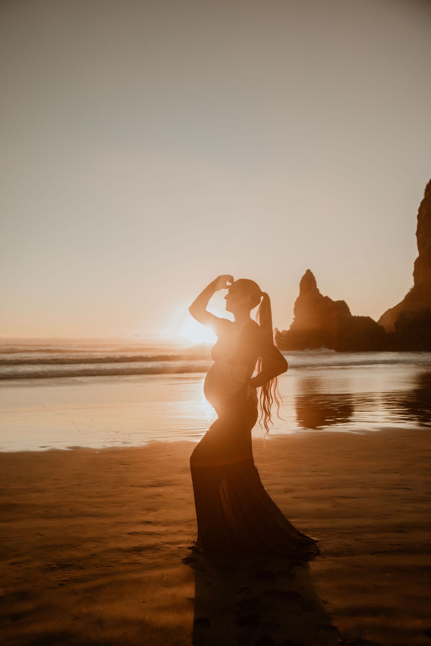 Silhouette of a pregnant woman posing on a beach at golden sunset with ocean waves and rock formations.