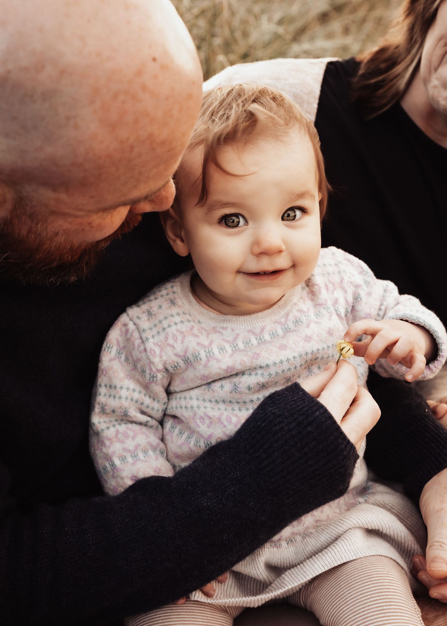 Adorable baby smiling at camera while being held by parents in a cozy knit sweater during a family portrait session.