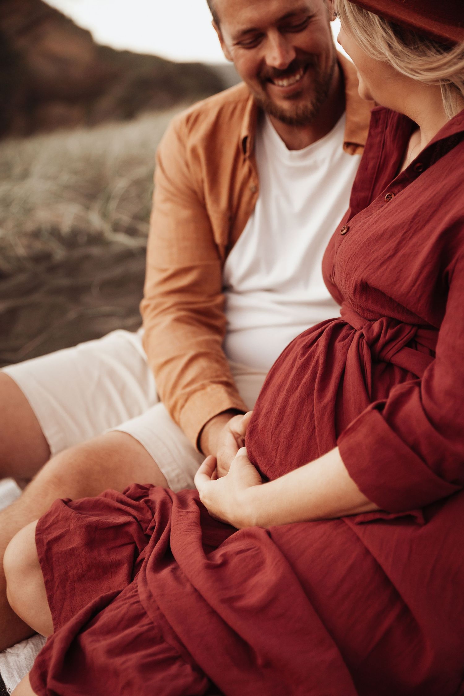 A smiling man in a brown shirt sits beside a pregnant woman in a burgundy dress, holding hands outdoors.