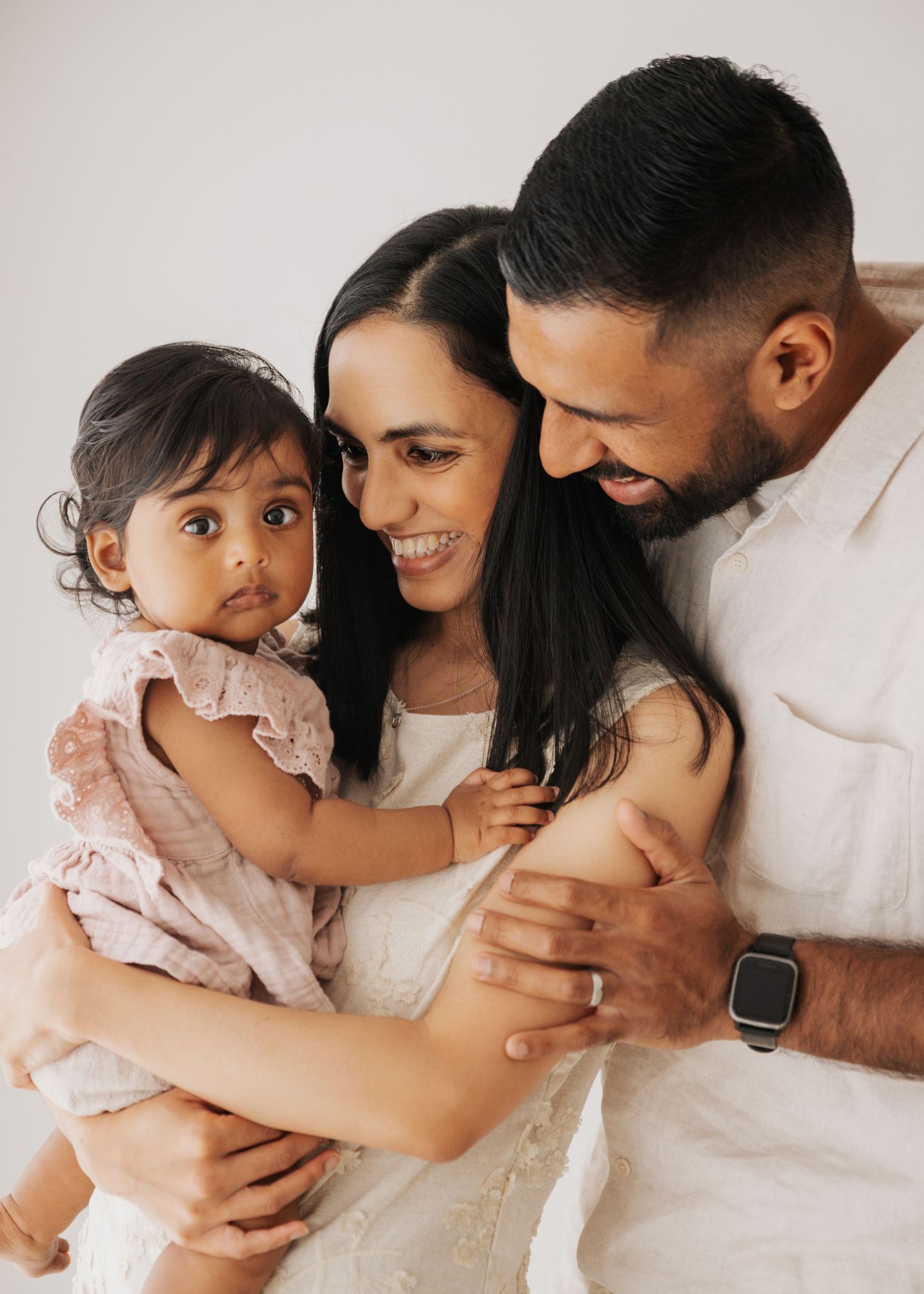 Smiling mother and father in white outfits lovingly embrace their baby daughter in a pink ruffled dress during a bright family portrait.