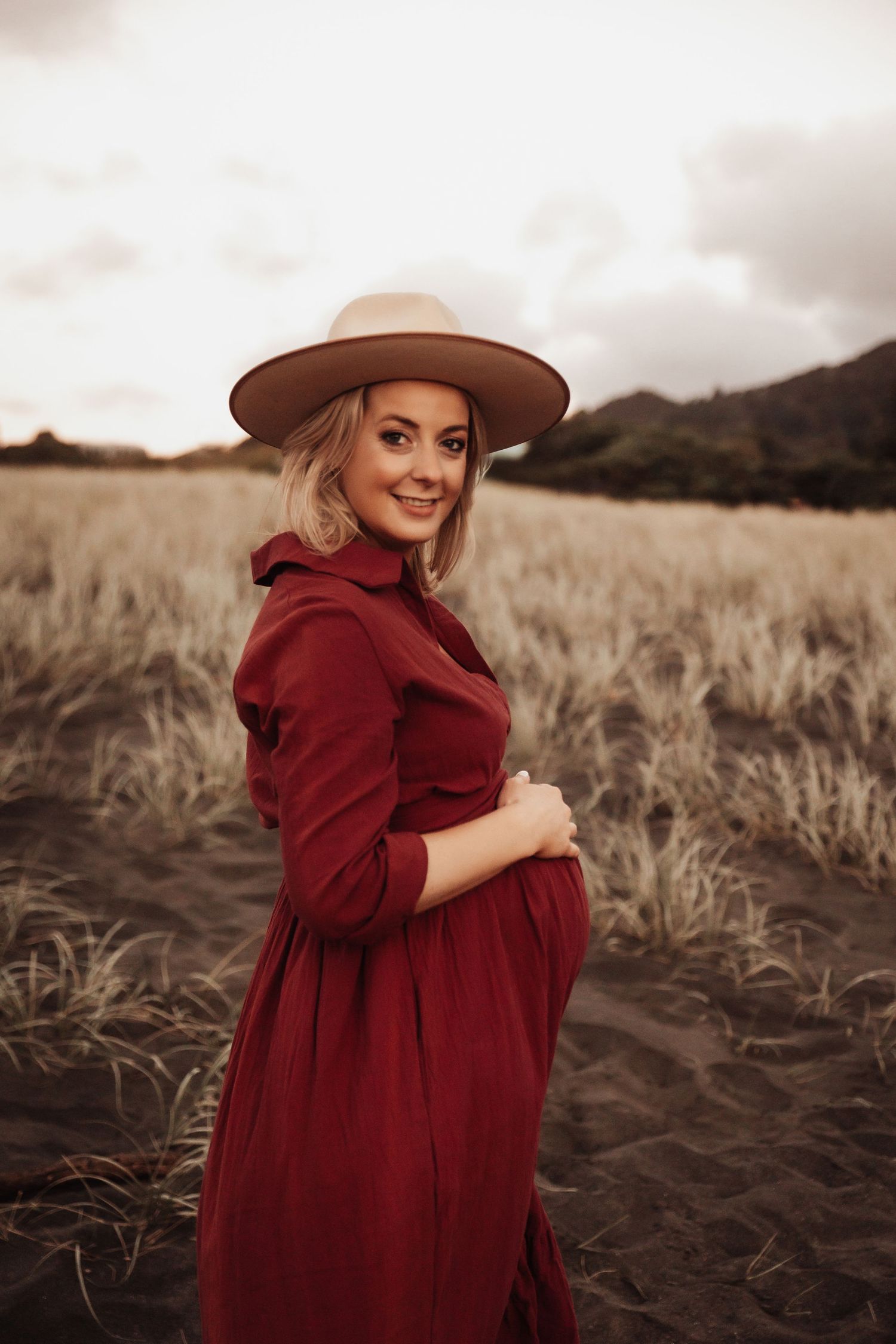 Pregnant woman in red dress and tan hat smiling in a golden field at dusk.