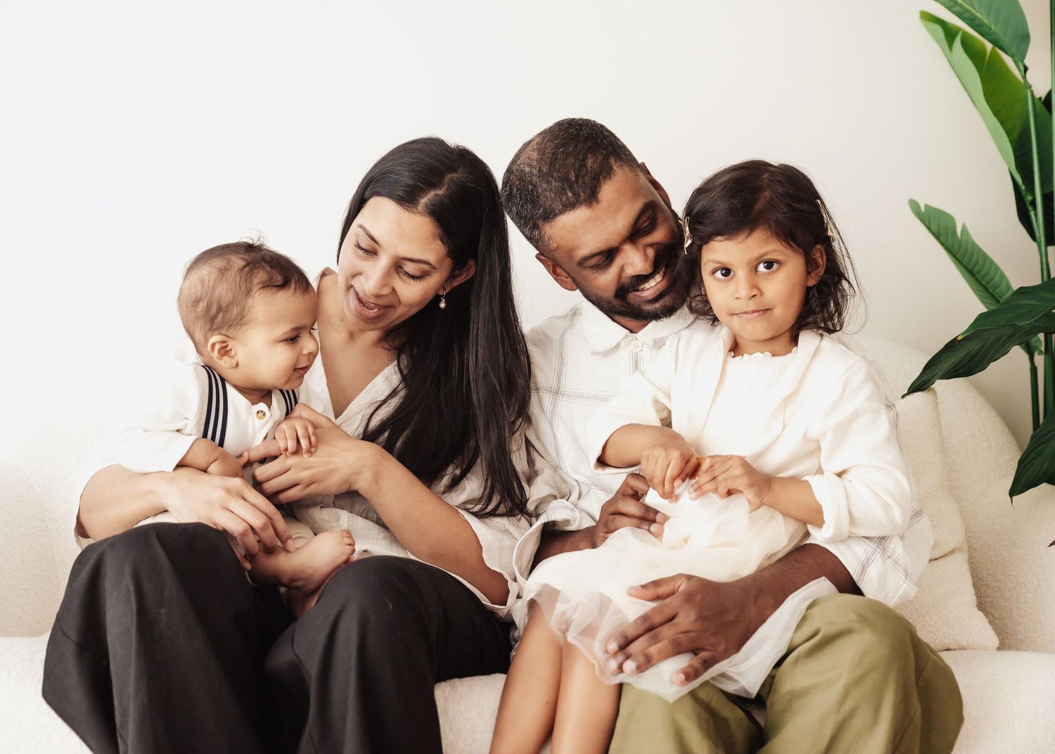 Happy family of four sitting on a white sofa, parents smiling with toddler and young child in a bright room.