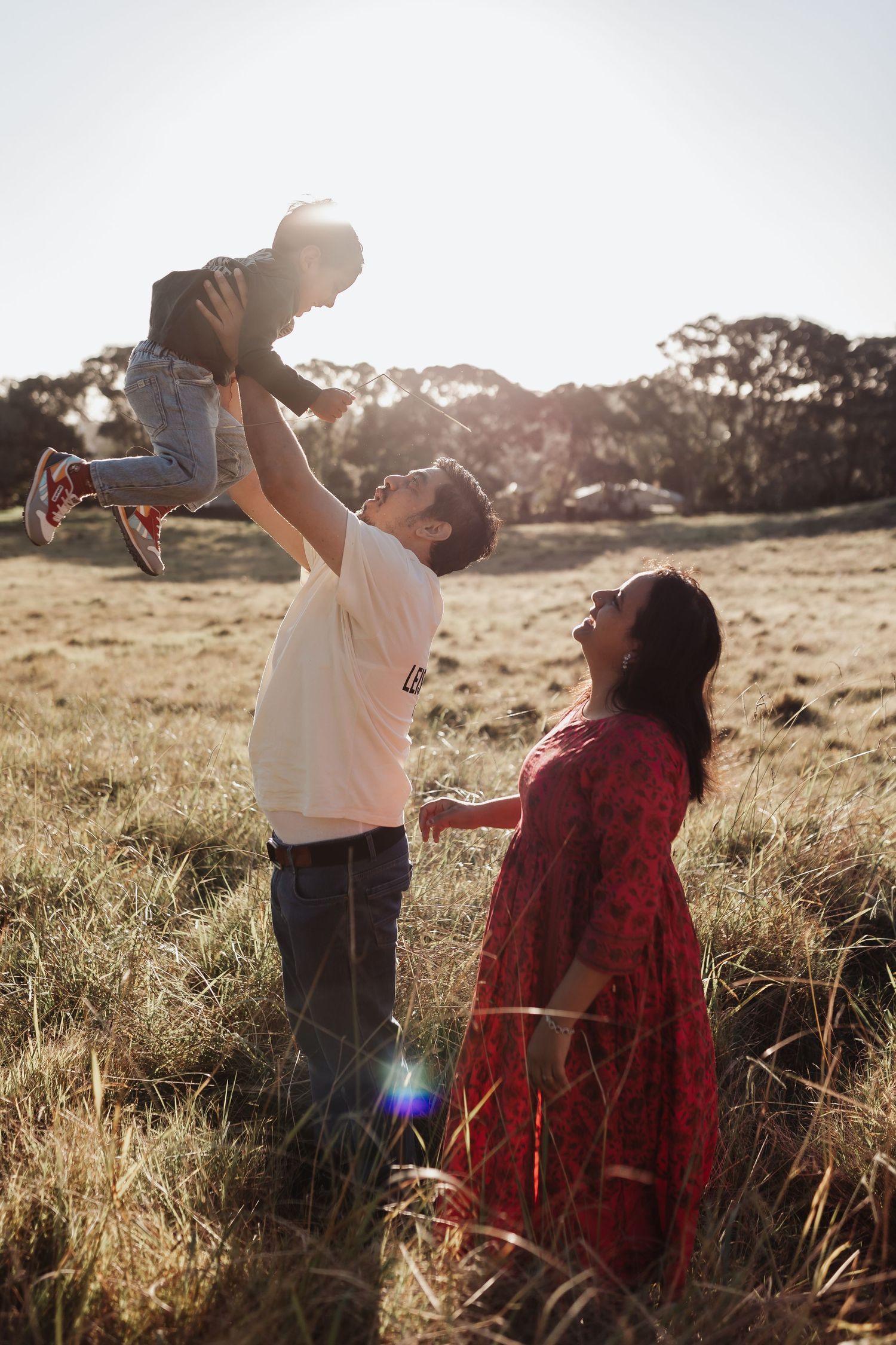 A joyful family in a sunny field, father lifting child in the air while pregnant mother watches and smiles.