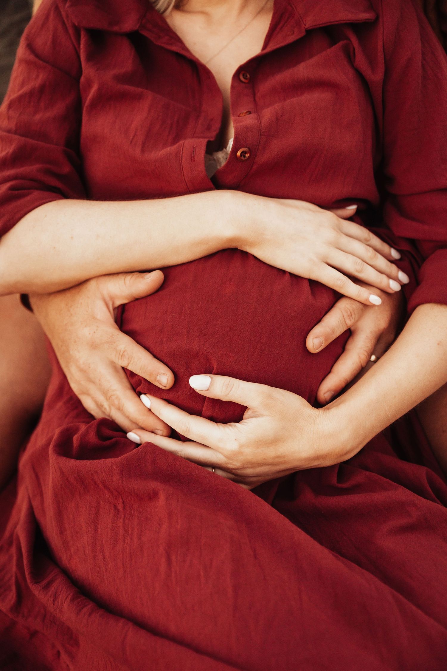 Pregnant woman in red dress with partner's hands forming heart shape on baby bump.