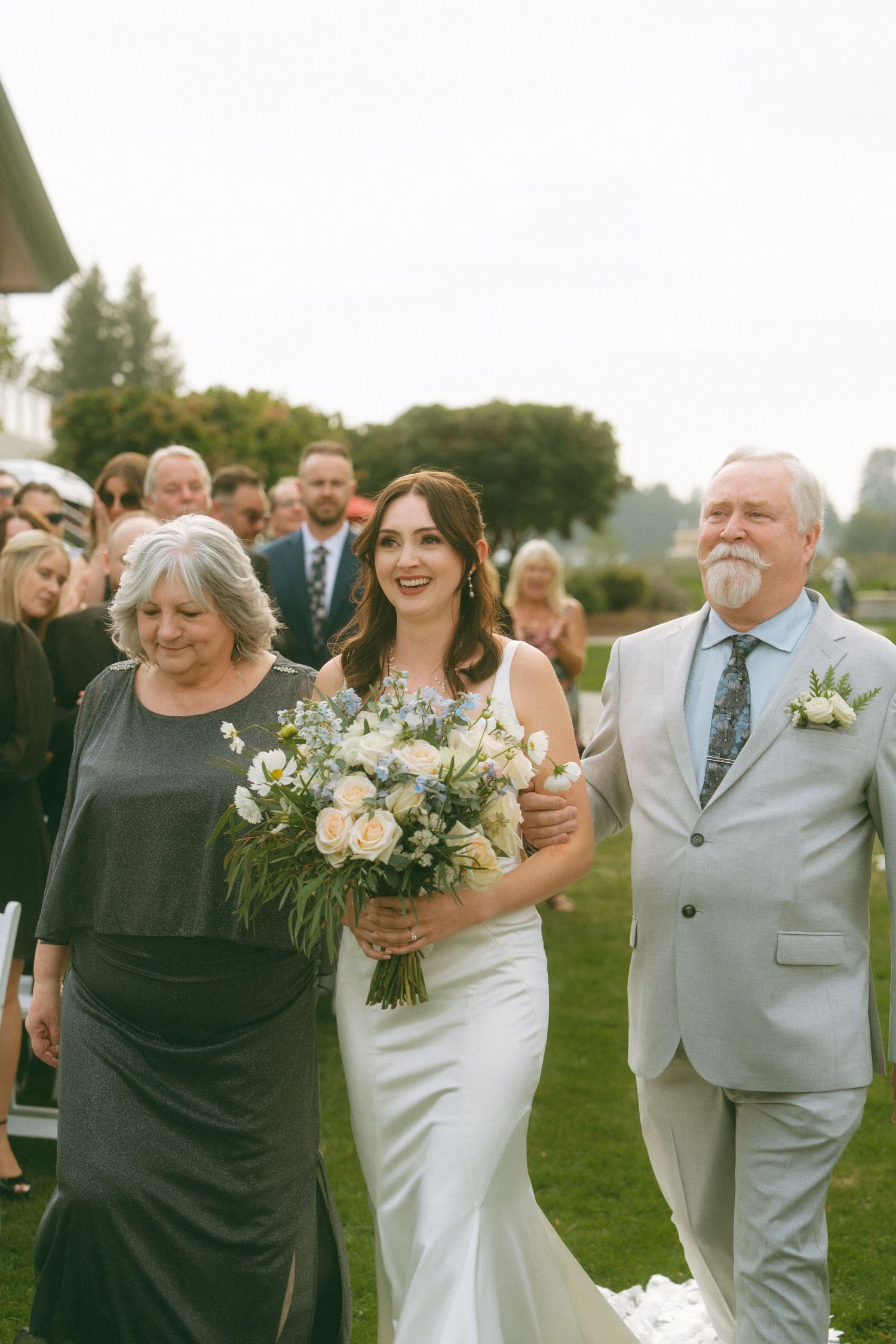 A bride walks down an outdoor aisle with her bouquet surrounded by wedding guests.