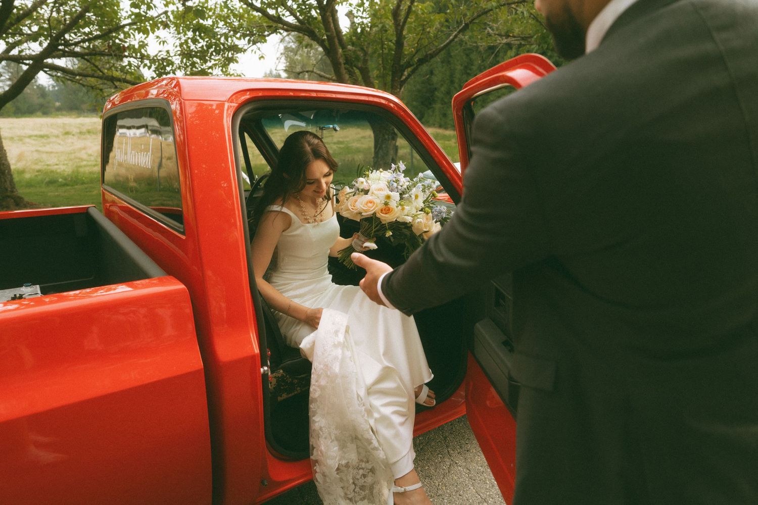 Wedding party gathering around a classic red vintage car on a tree-lined street.