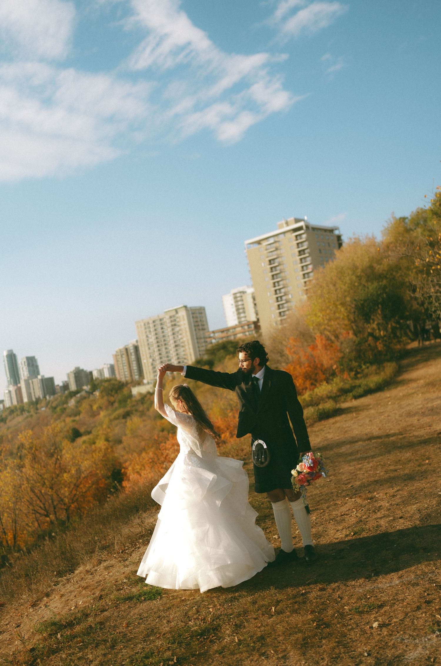 A couple poses for wedding photos against an urban autumn landscape with high-rise buildings.