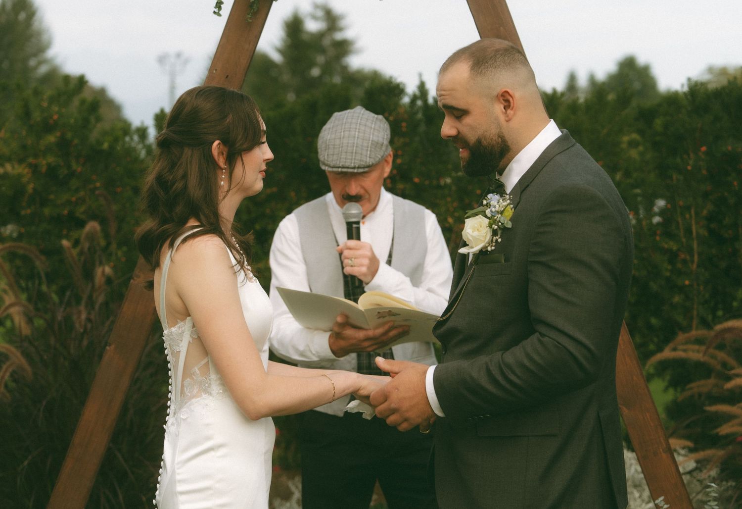 An outdoor wedding ceremony under a wooden arch with an officiant.