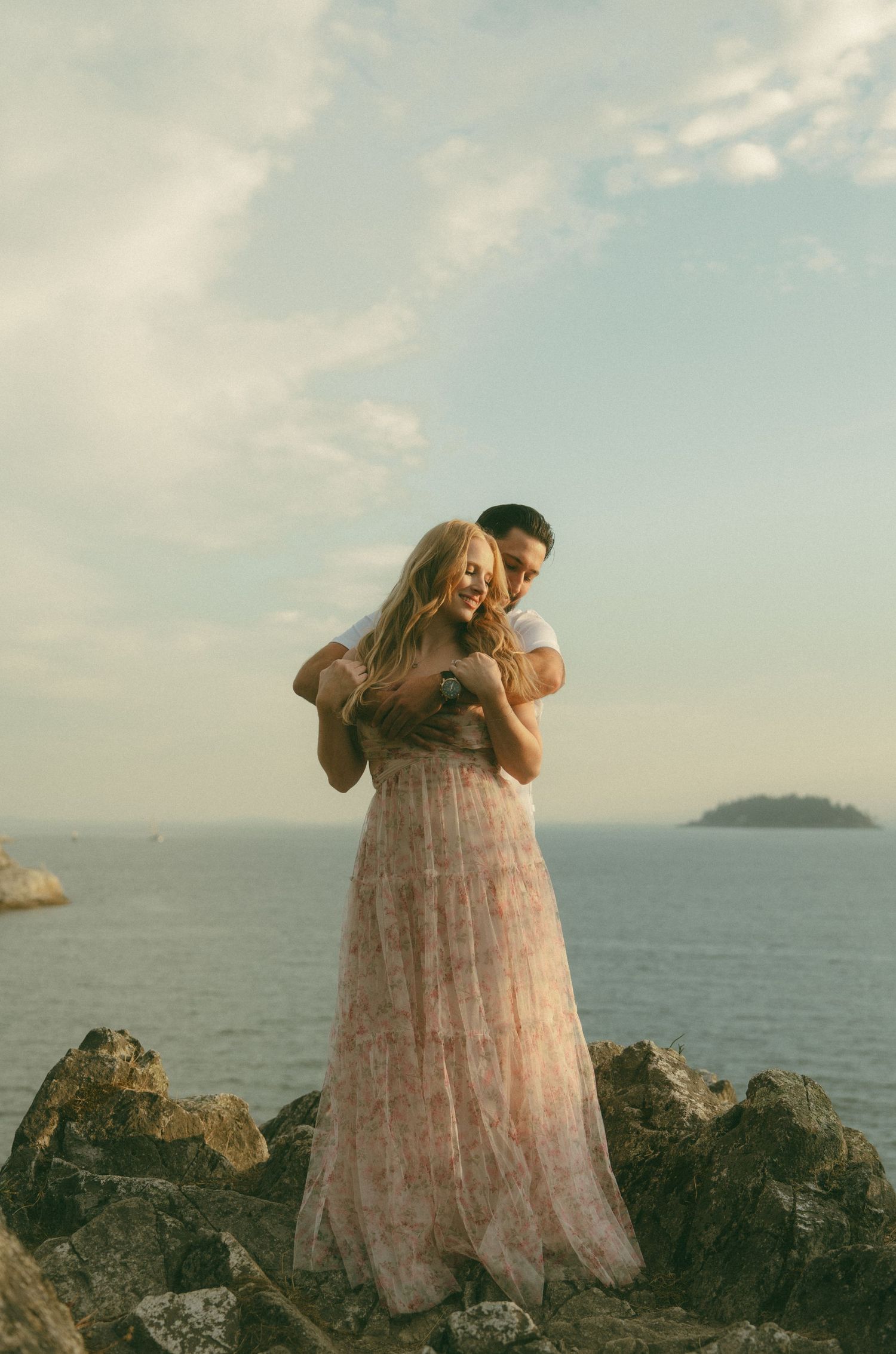 Romantic couple embraces against coastal backdrop in blush pink formal attire.