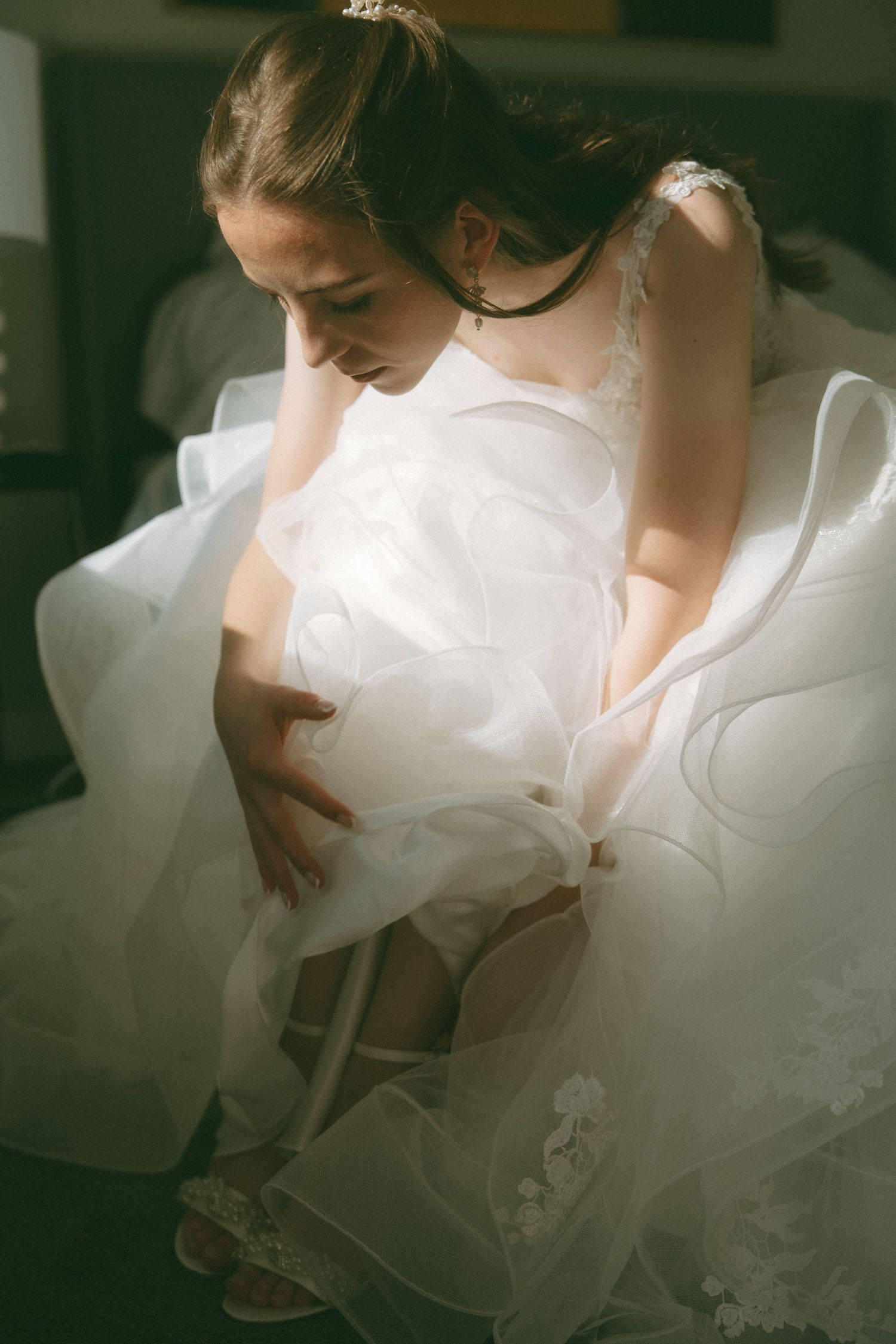 A bride in a flowing white wedding dress adjusts her gown in soft natural lighting.