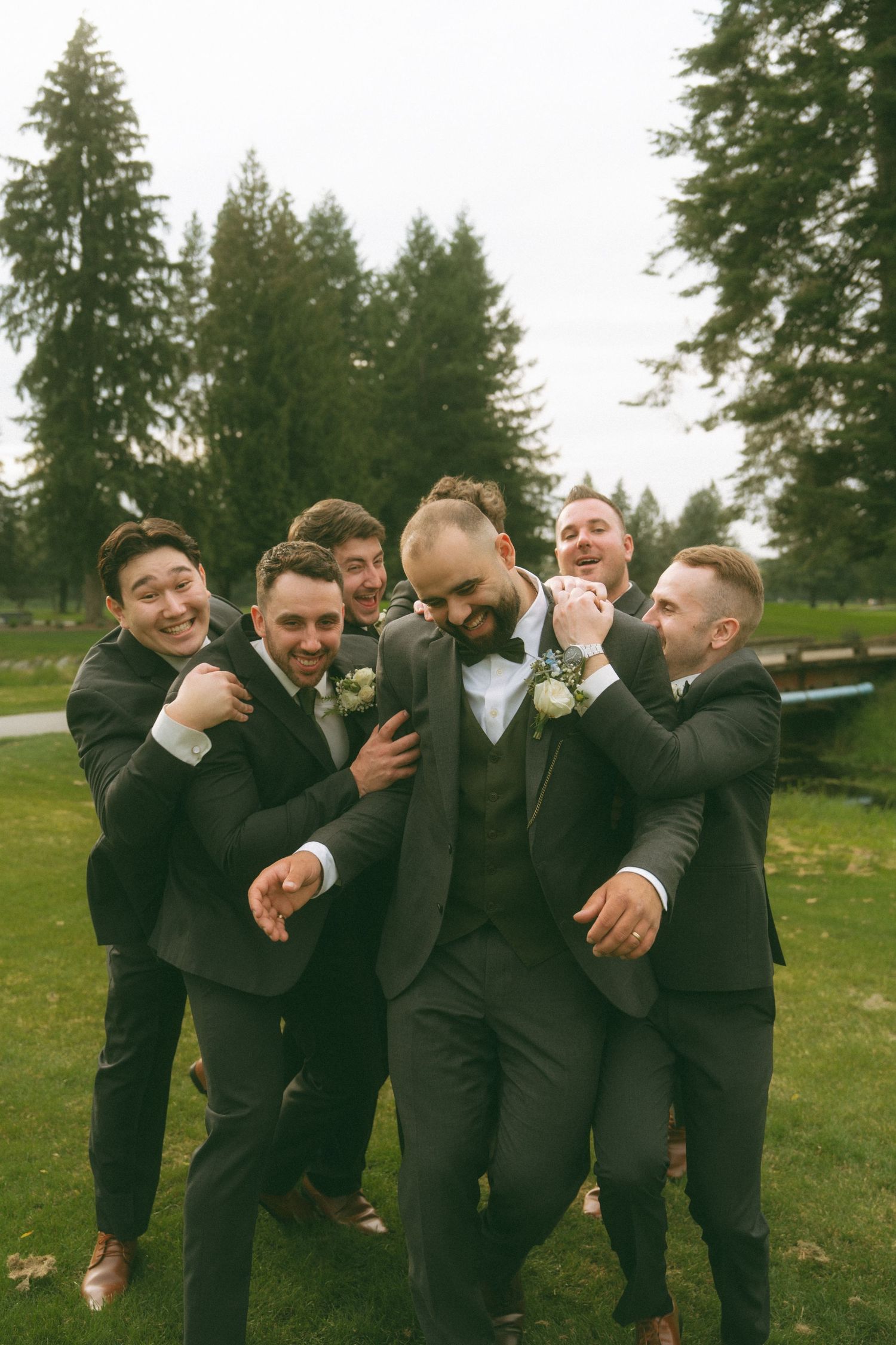 Groomsmen pose playfully in a line wearing black suits outdoors.