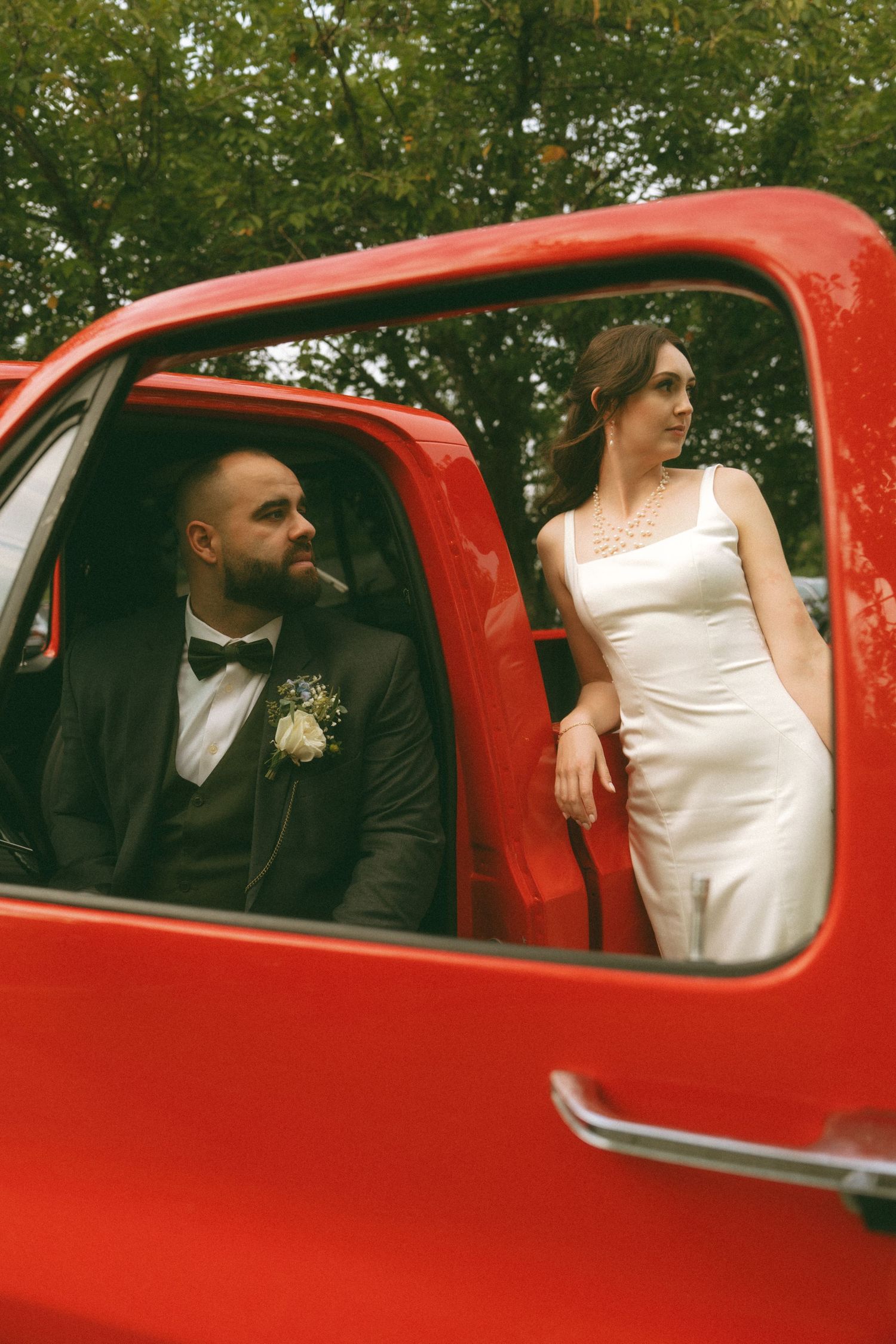 Wedding couple poses inside vintage red car for artistic photo series.