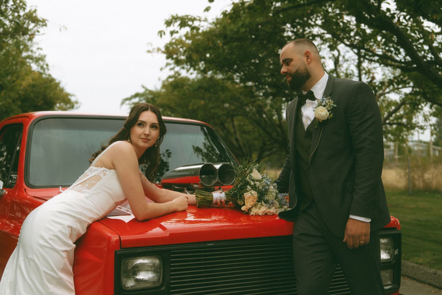 Couple poses with vintage red car during wedding photoshoot.