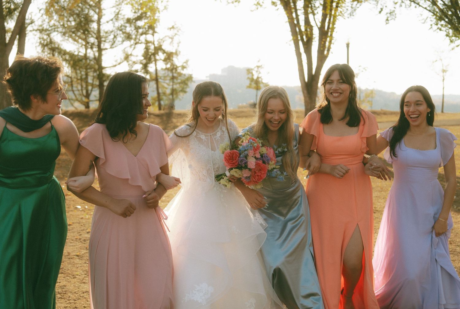 Bridesmaids in colorful dresses walk together outdoors during golden hour.