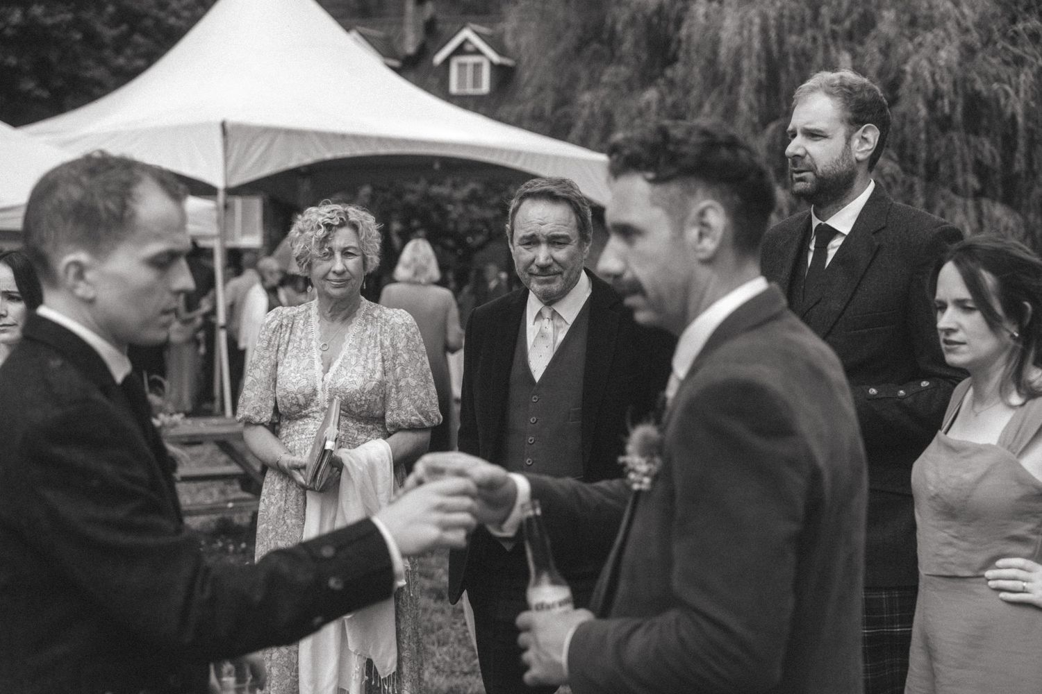 Wedding guests enjoying drinks and conversation under a tent at an outdoor celebration.