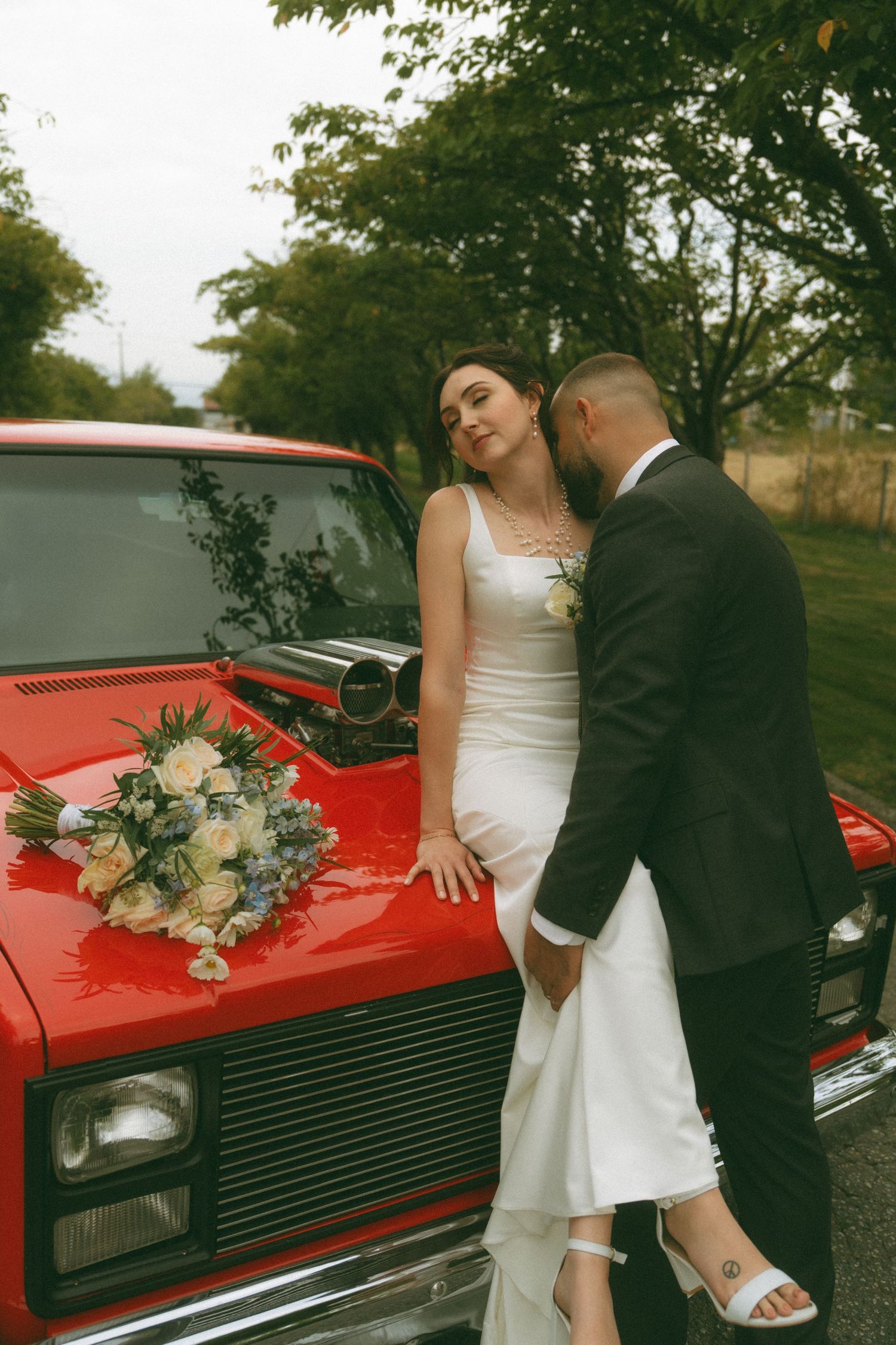 Newlyweds share intimate moment against classic red car during outdoor photo session.